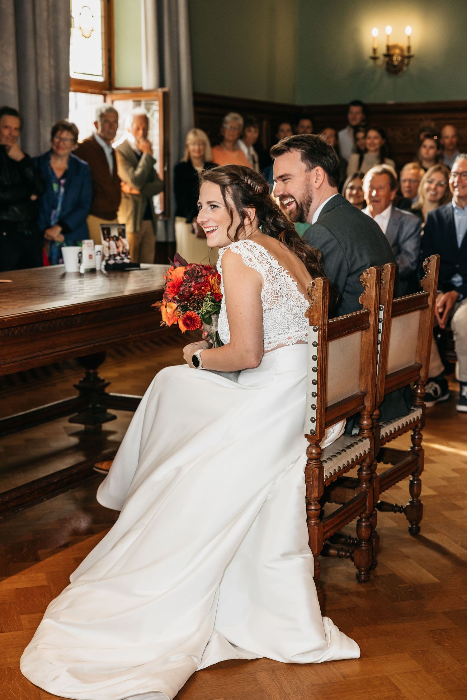 Bride in white lace gown holding red bouquet seated with laughing groom at civil wedding ceremony.