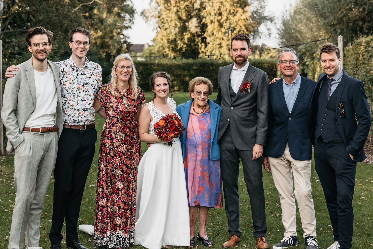 Bride and groom with six family members posing for formal wedding portrait on green lawn with autumn foliage.
