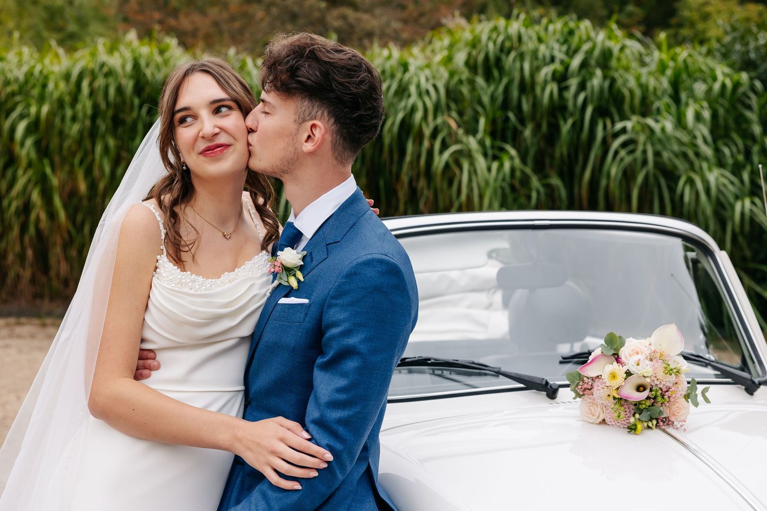 Groom in blue suit kissing bride on cheek beside a classic white VW Beetle convertible.