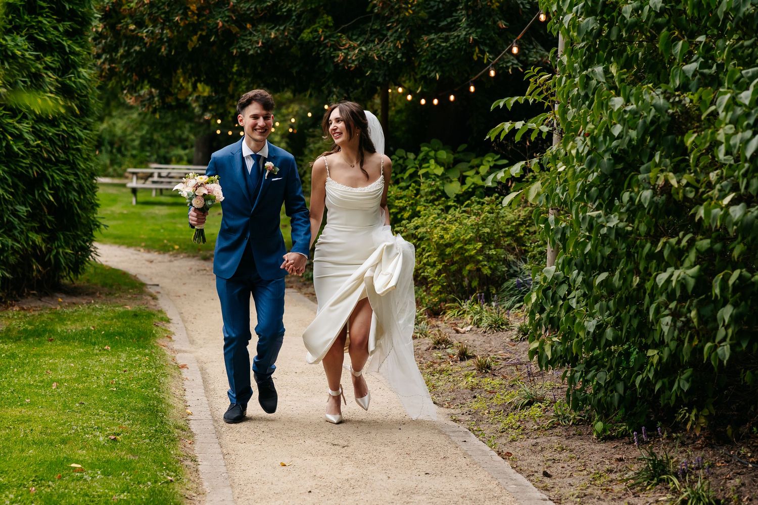 Bride and groom walking joyfully along a lush garden path, bride holding her dress, groom in a blue suit carrying flowers.
