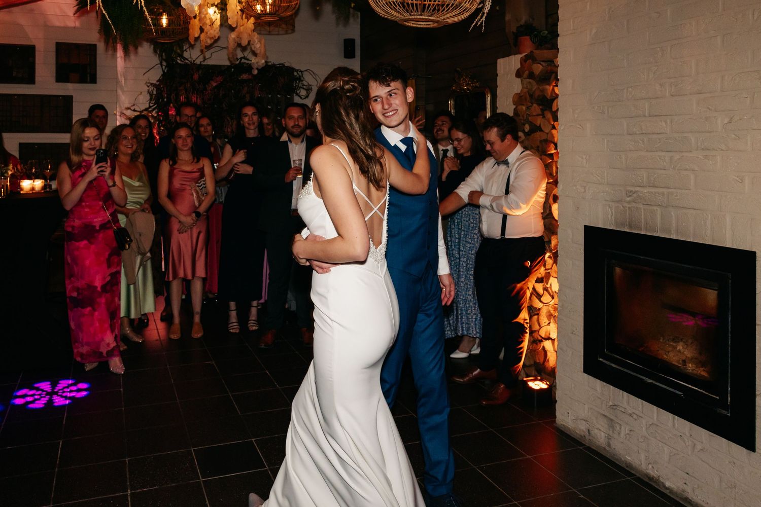 Bride and groom share romantic first dance near glowing fireplace while wedding guests look on and celebrate.