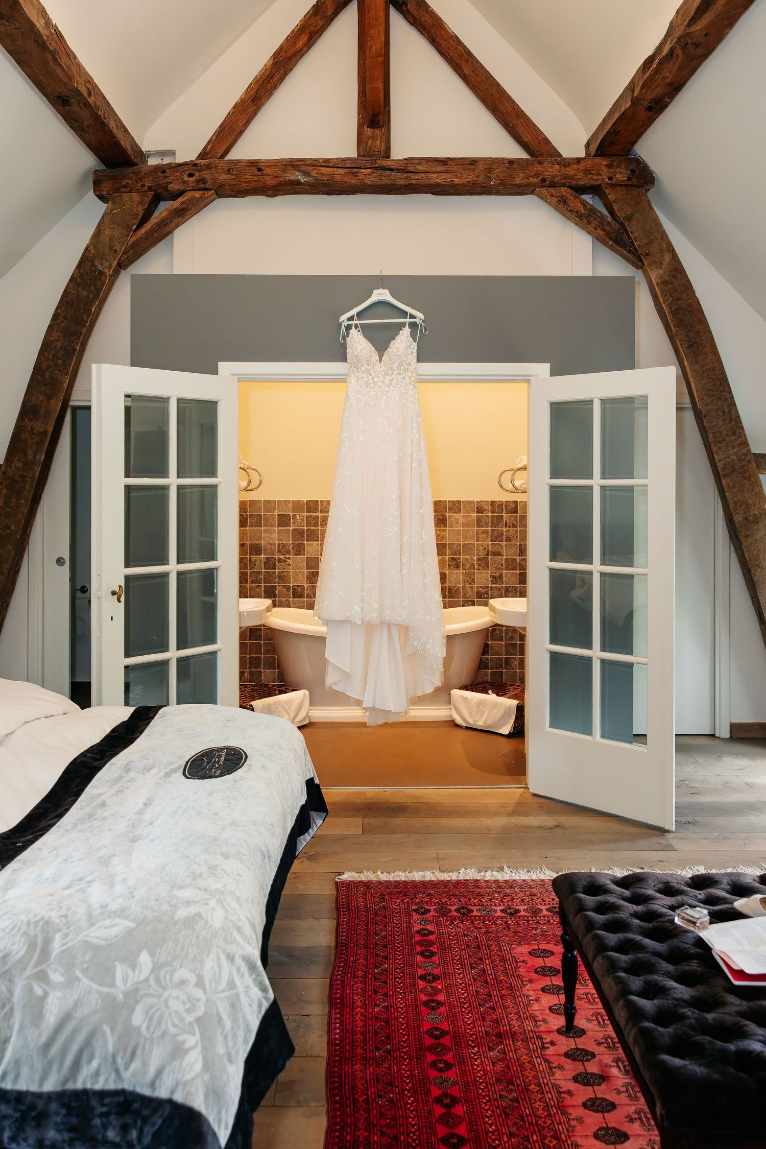 Wedding dress hanging in rustic attic bedroom with exposed wooden beams and French doors.