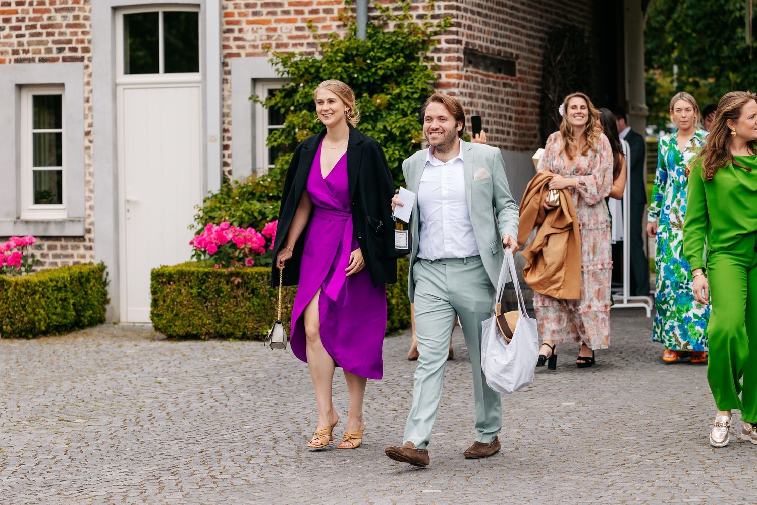 Smiling wedding guests in colorful attire walk together across a cobblestone courtyard.