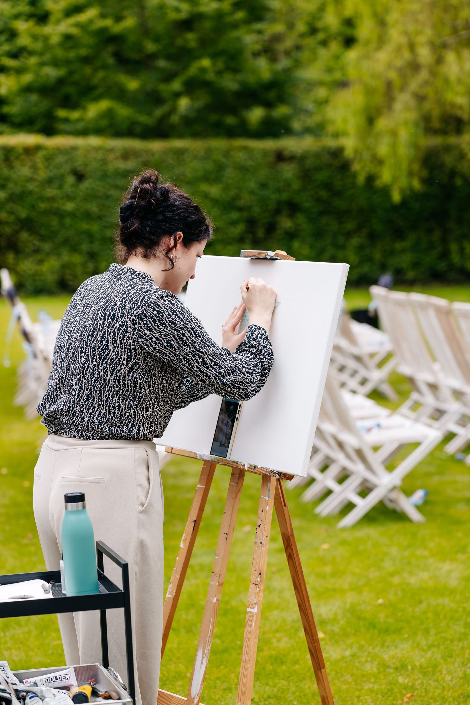 Artist painting on canvas at easel during outdoor wedding reception with white chairs on green lawn.