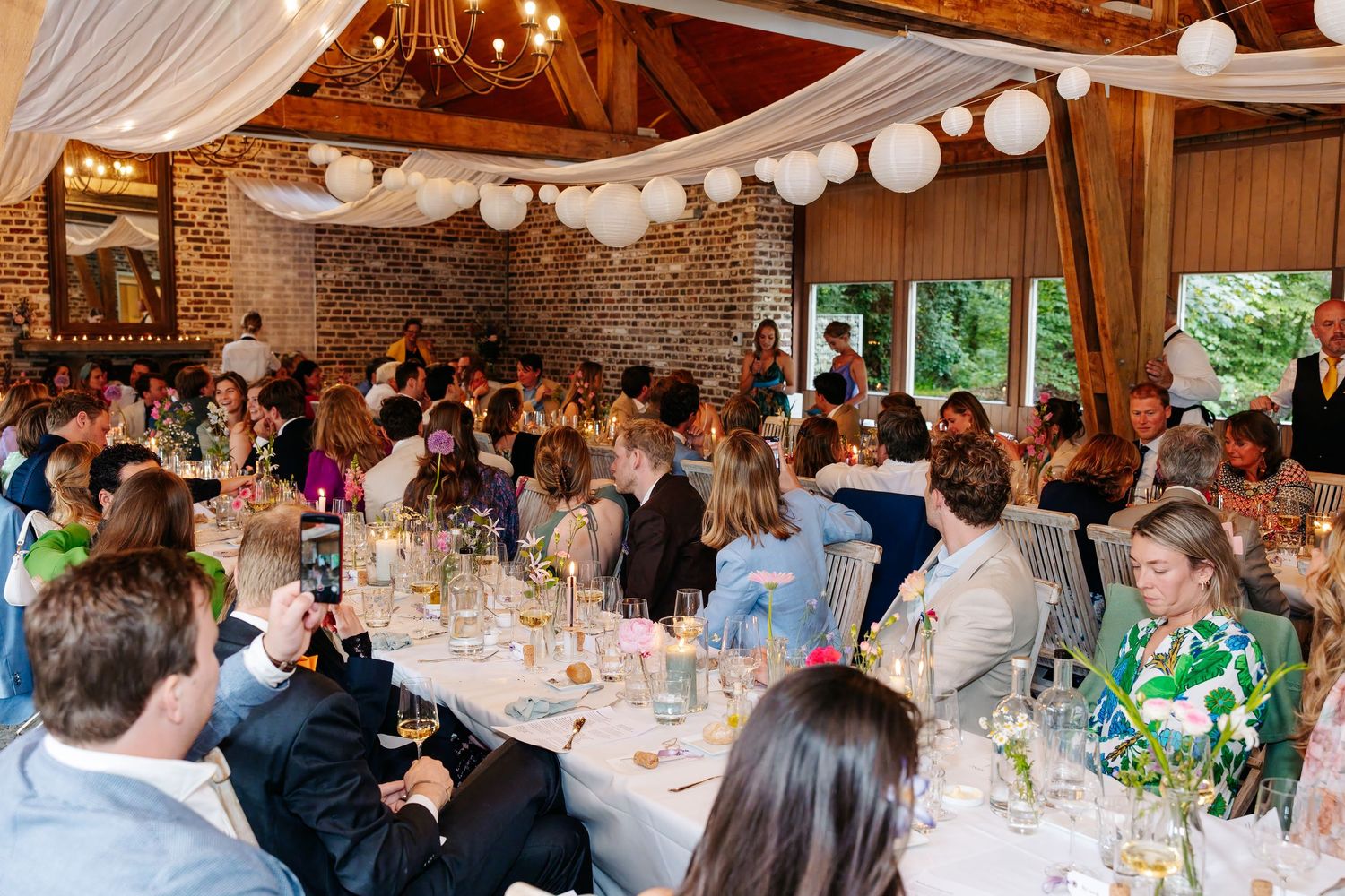 Large wedding reception crowd seated at decorated tables inside rustic brick venue with white balloon ceiling.