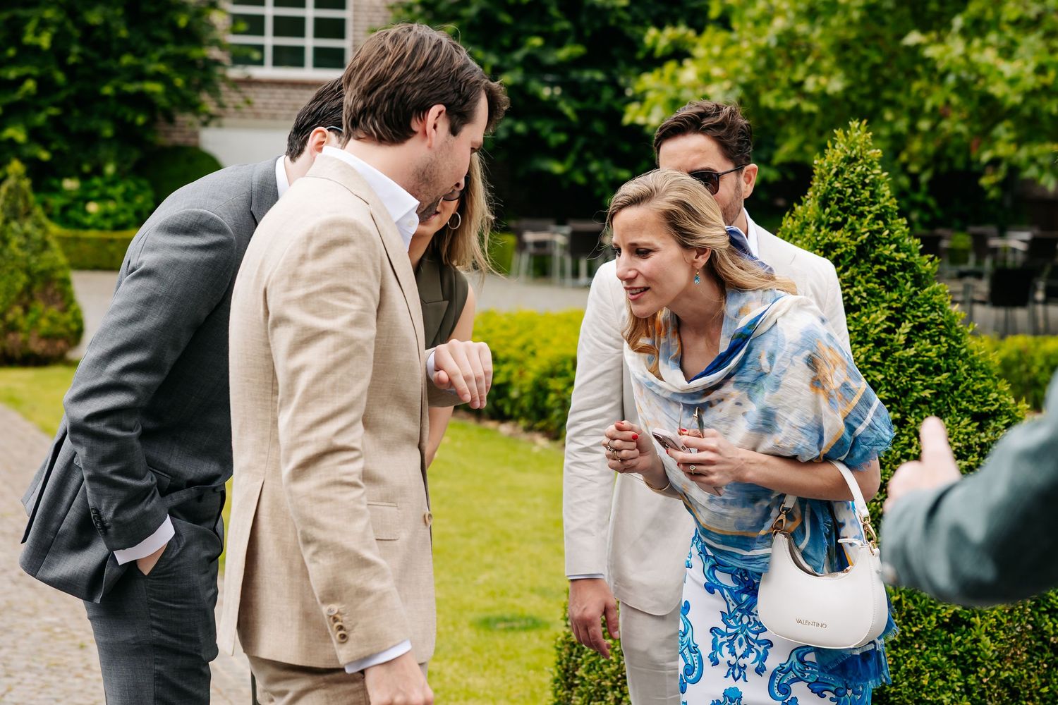 Wedding guests in elegant attire socializing outdoors in a lush green garden during a summer wedding celebration.