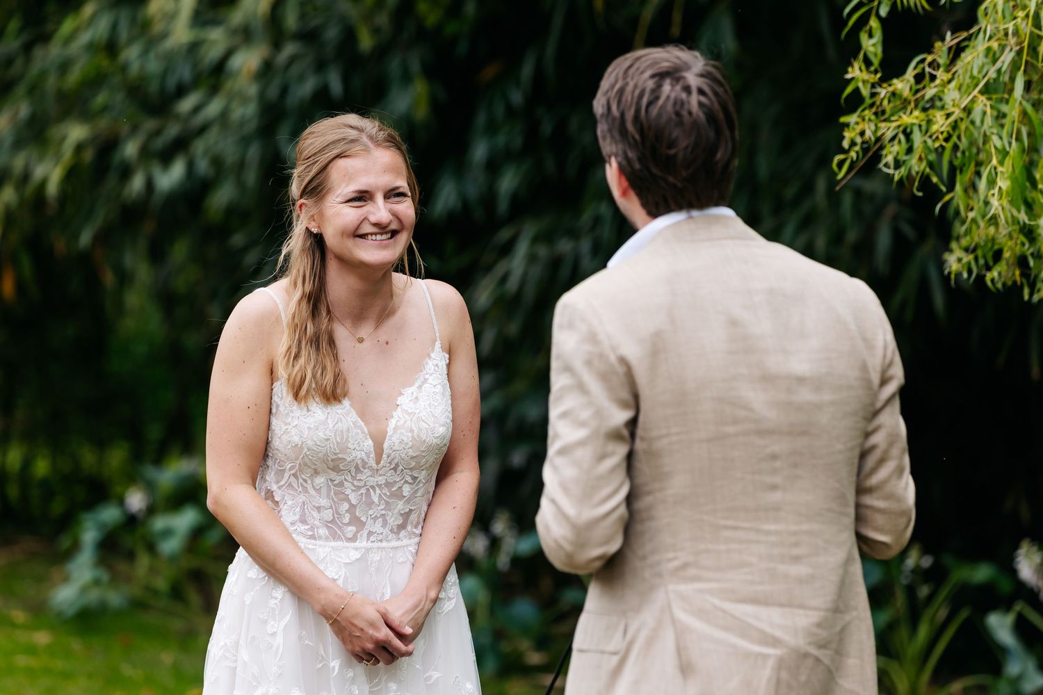 Bride in lace spaghetti strap wedding dress smiling joyfully at groom during outdoor garden wedding ceremony.