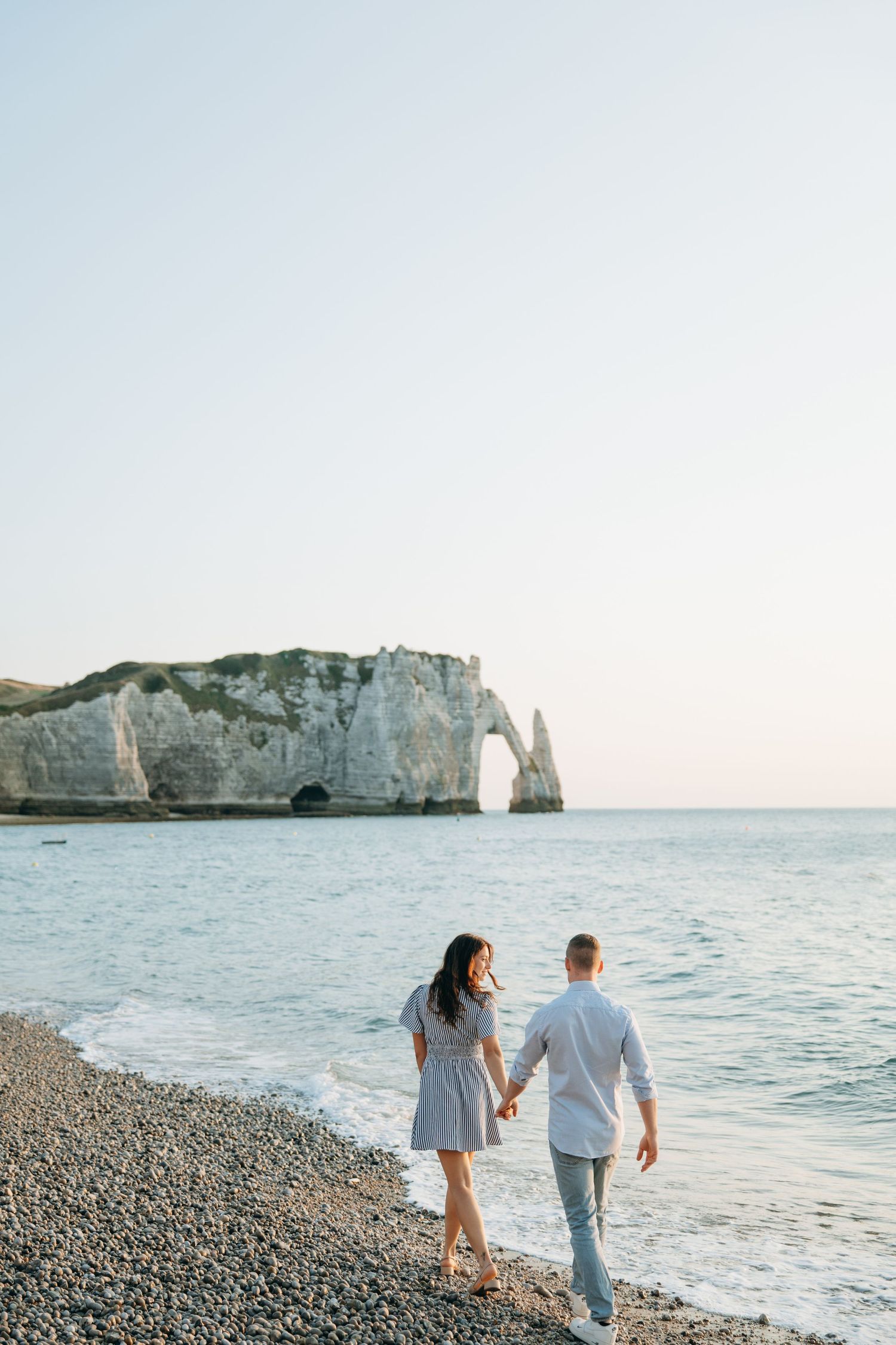Couple walking hand in hand on pebble beach with stunning white chalk cliffs and sea arch backdrop.