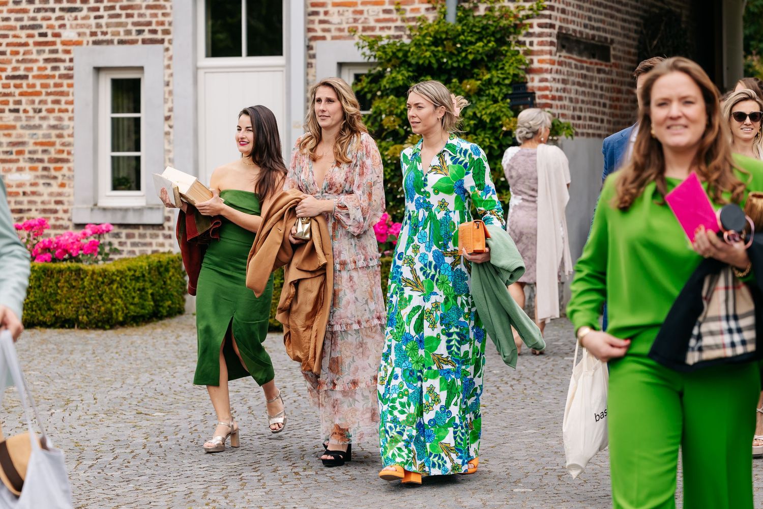 Wedding guests in colorful summer dresses walk across cobblestone courtyard at outdoor venue.