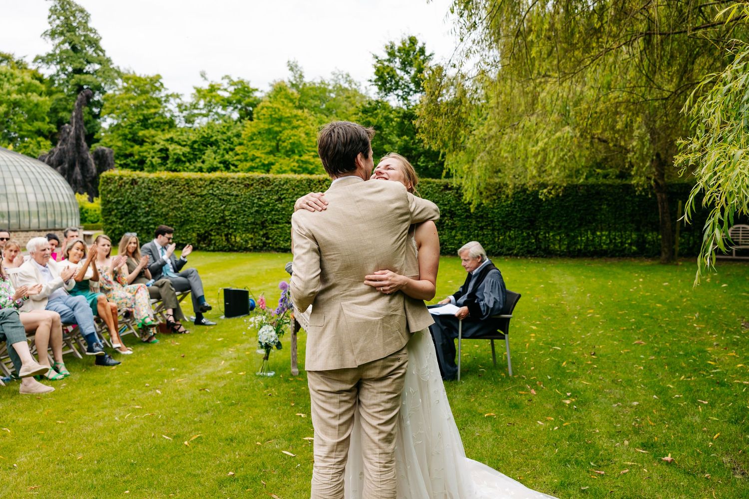 Bride and groom embrace during outdoor wedding ceremony while guests watch seated on chairs in lush green garden.