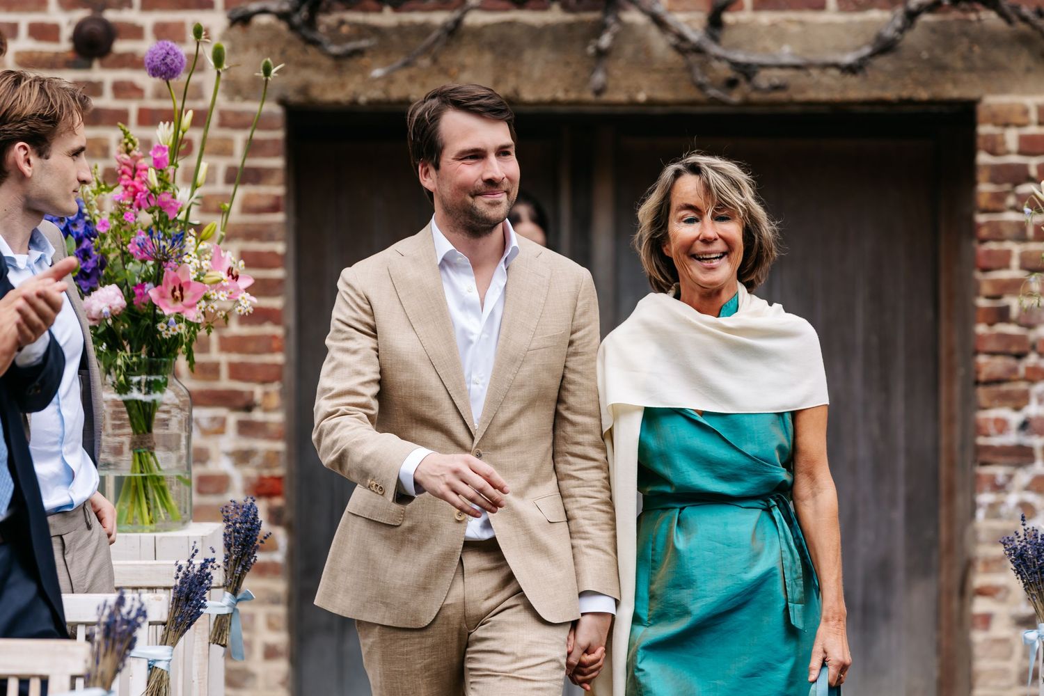 Groom in tan suit escorts elegantly dressed woman in teal dress at outdoor wedding ceremony.