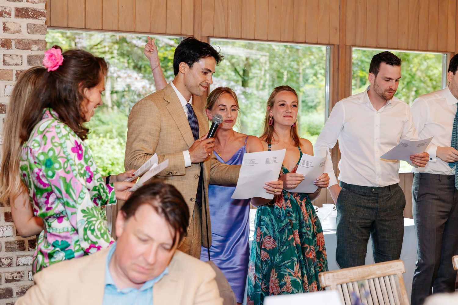 Wedding guests mingle and review programs in an elegant venue with large windows and natural light.