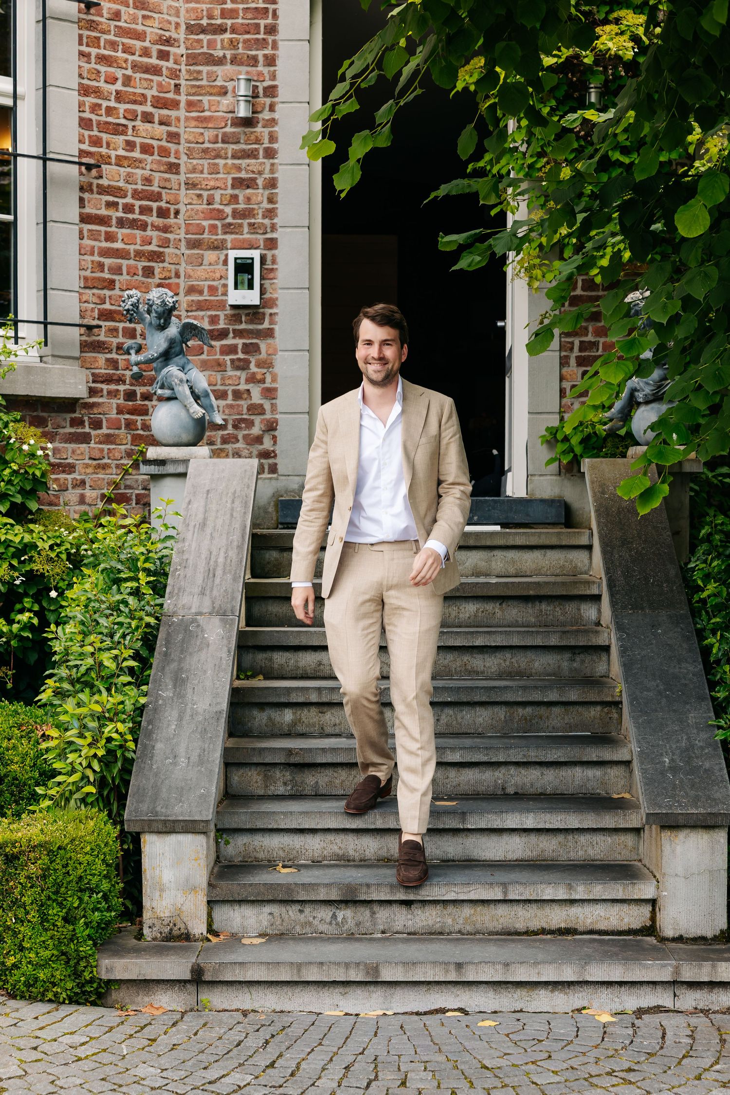 Smiling man in beige suit walking down stone steps of elegant brick mansion with garden.