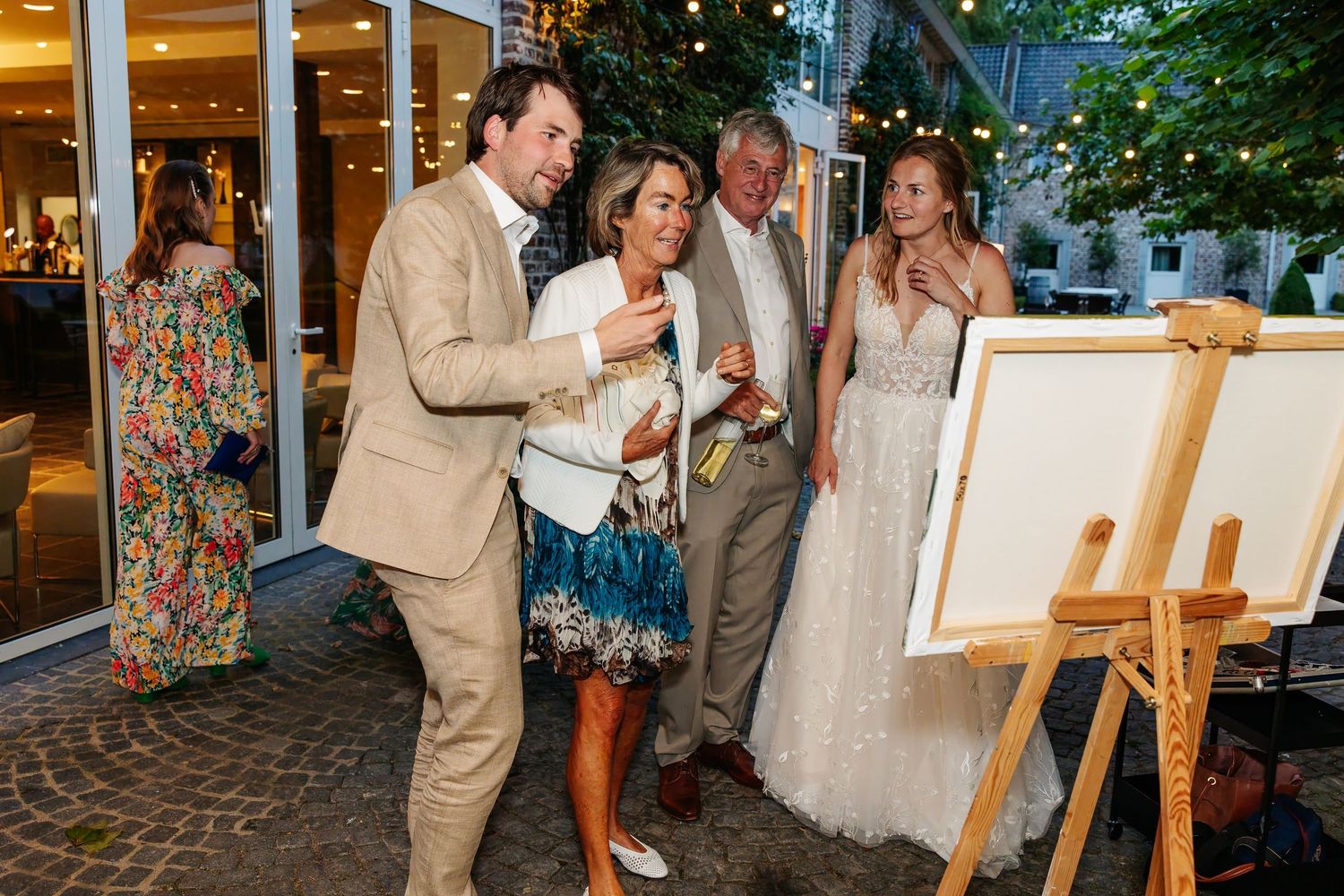 Bride in white gown and guests admire artwork on easels at an elegant outdoor wedding reception.