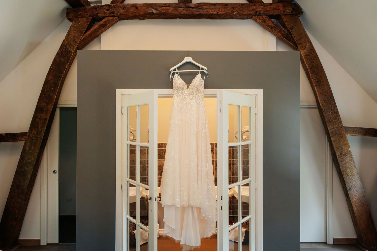 Delicate bridal gown displayed on hanger above white French doors beneath rustic exposed wooden beams.
