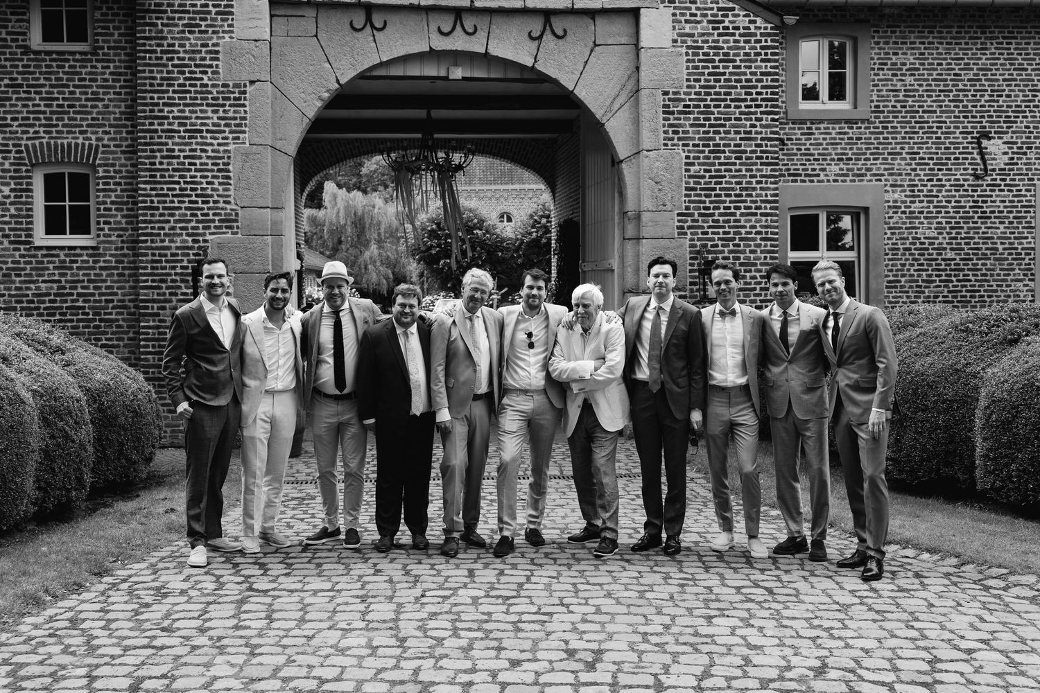 Black and white group portrait of groomsmen in suits posing at brick archway courtyard.