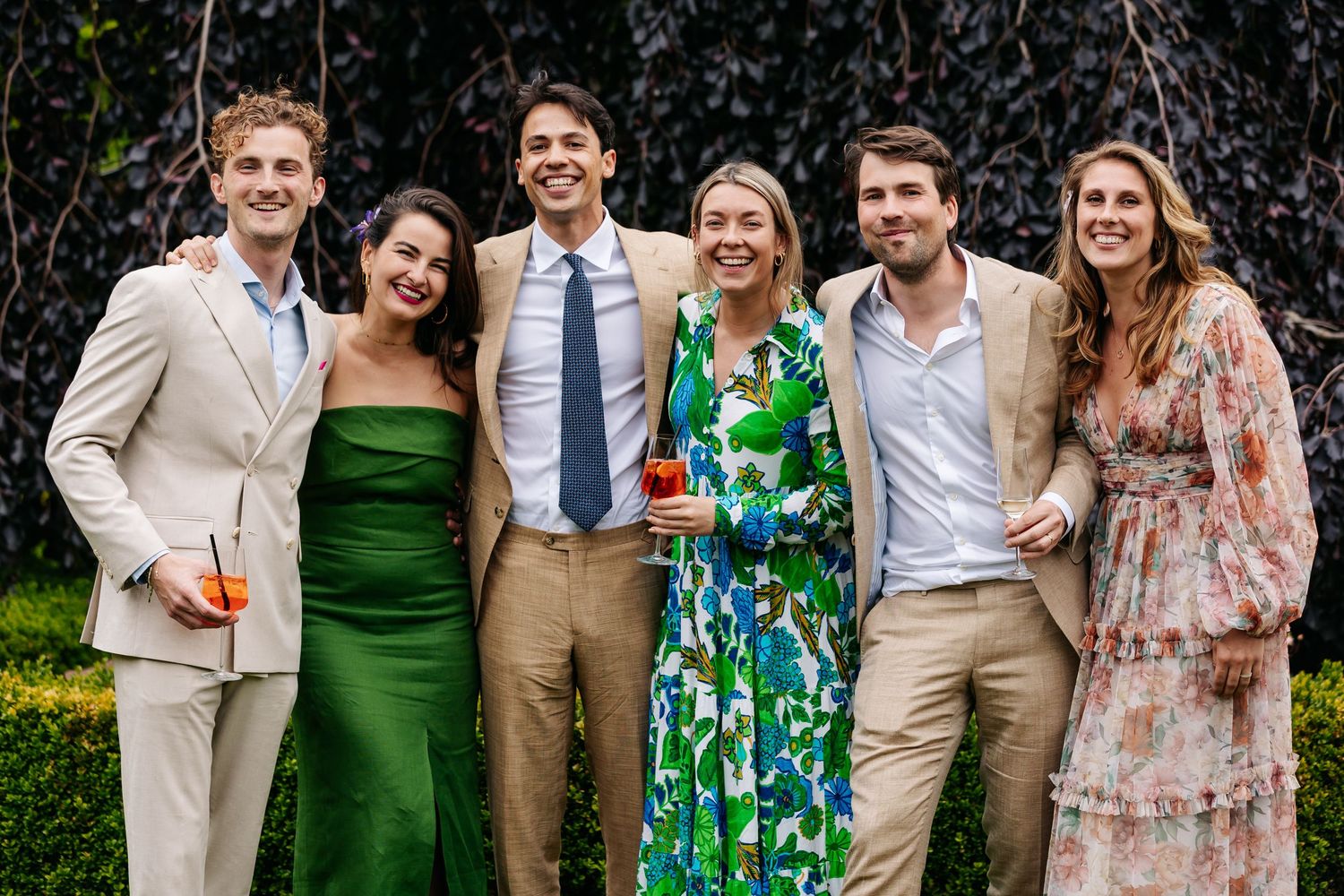 Group of six elegantly dressed friends smiling together at an outdoor wedding reception.