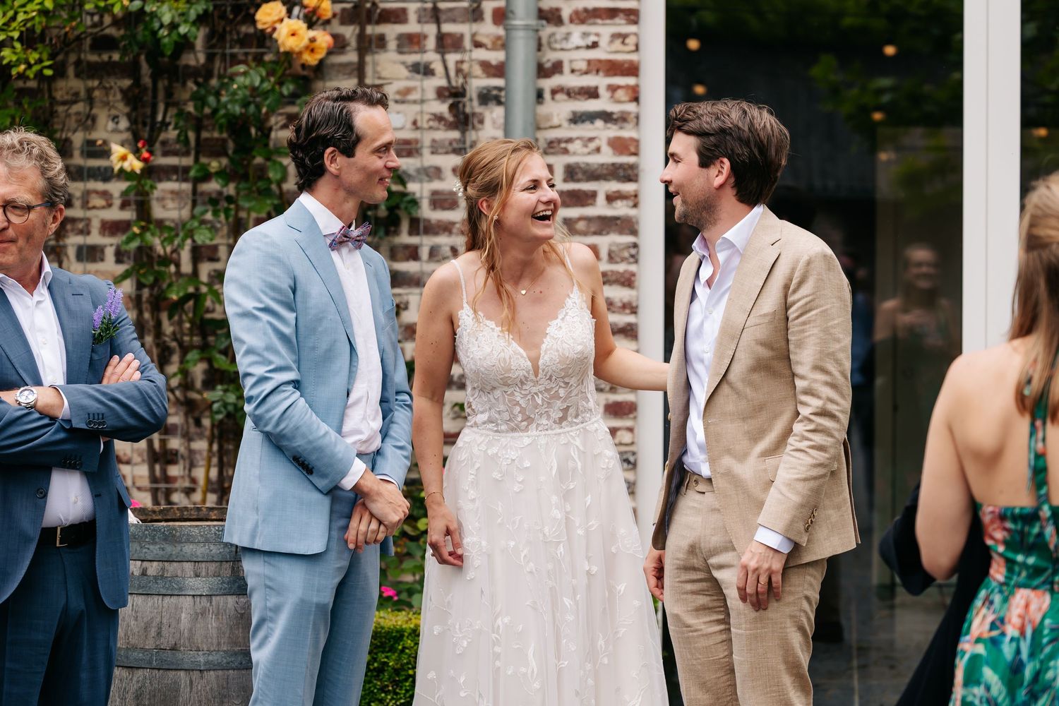 A laughing bride in a lace gown stands between two suited men at an outdoor wedding.