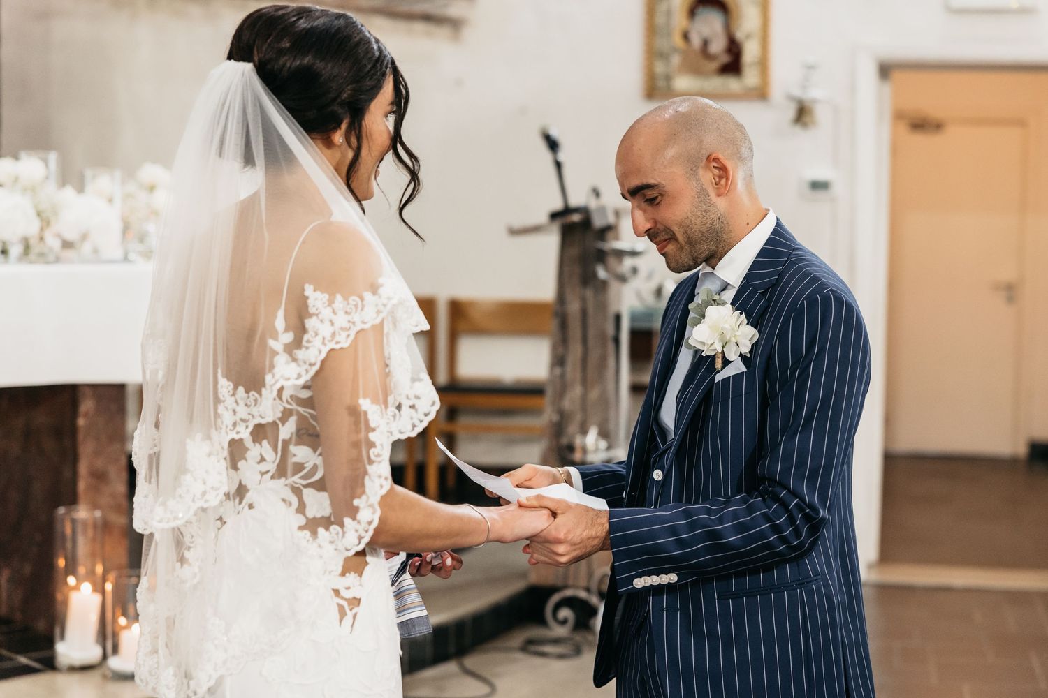 Groom in navy suit exchanges rings with bride in lace dress at church altar.