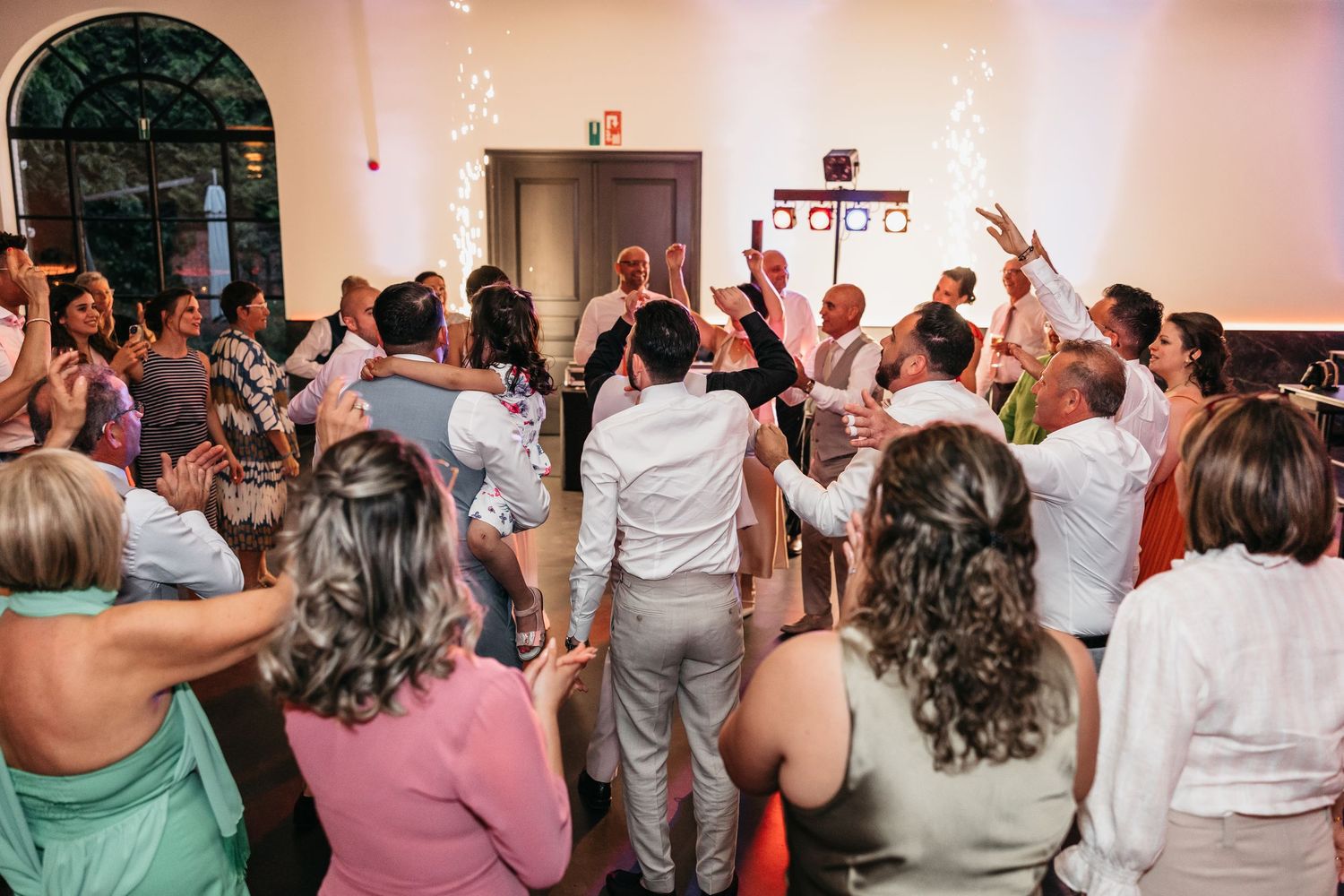 Wedding guests surrounding groom on packed dance floor during lively reception celebration.