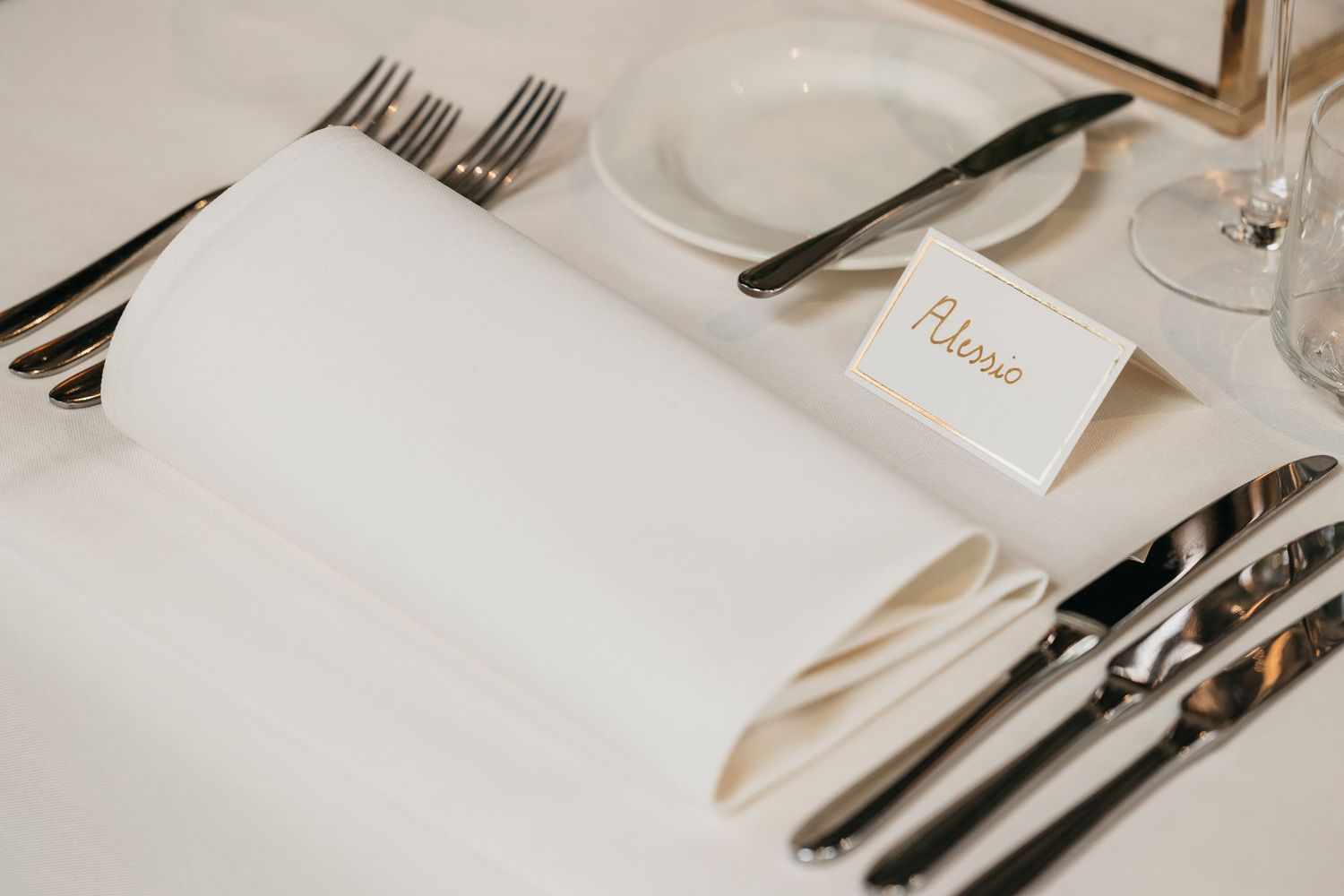 Wedding place setting with white folded napkin and a handwritten name card reading Adiano on white tablecloth.