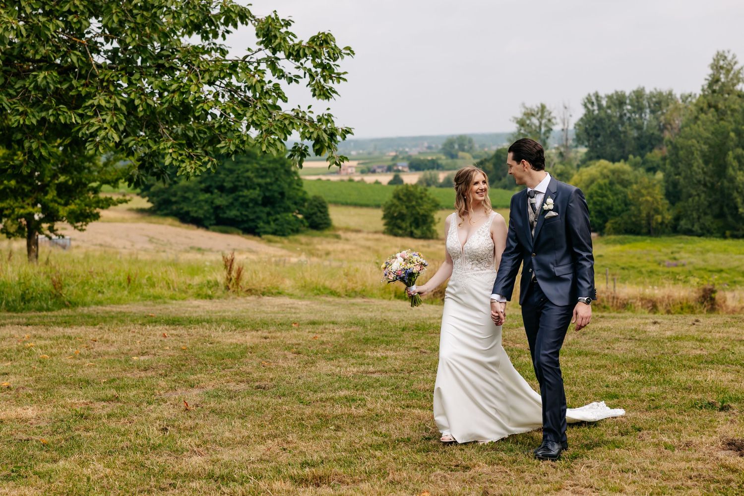 Smiling bride and groom strolling through a lush green meadow on their wedding day.