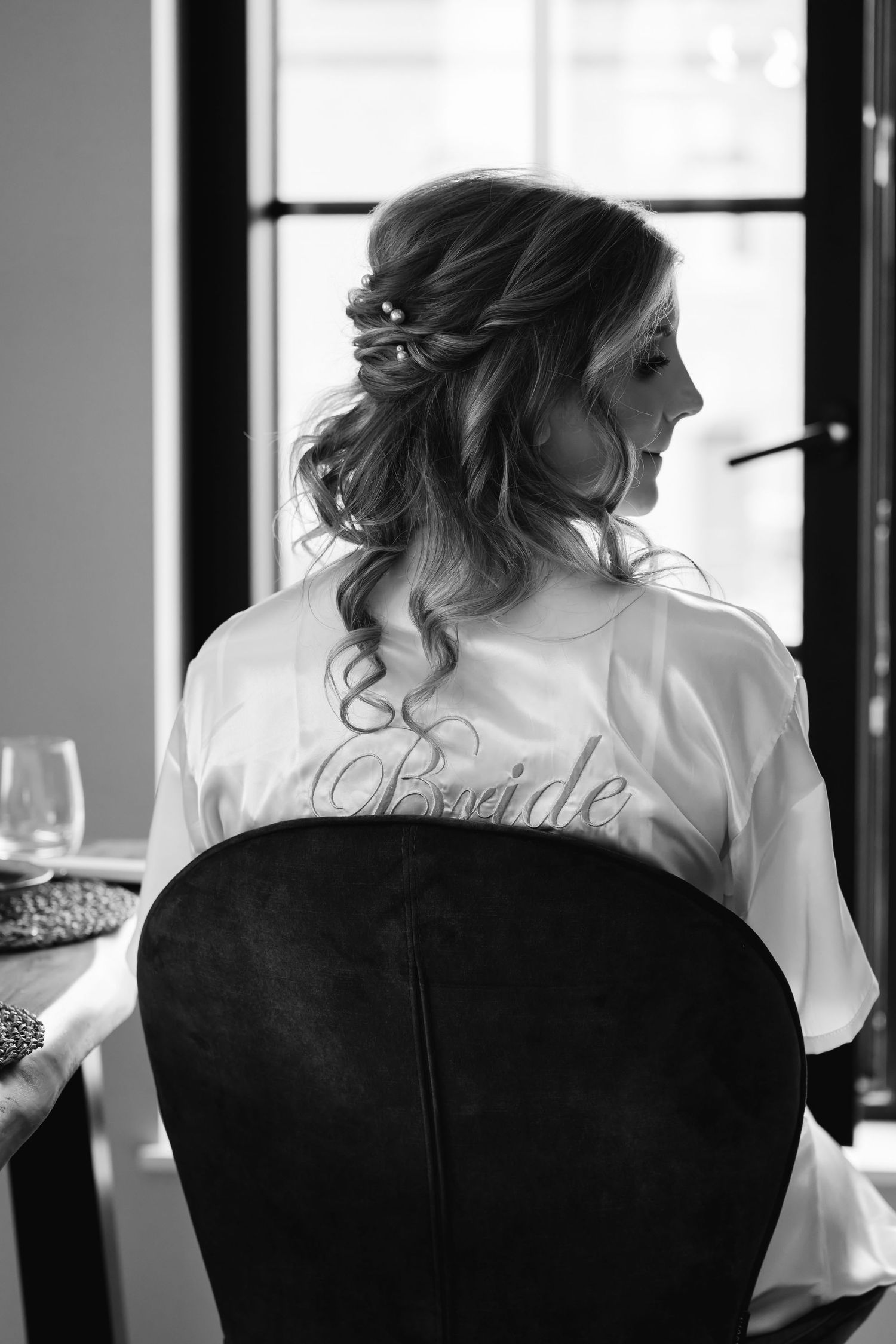Black and white photo of bride in Bride robe with elegant updo hairstyle seated by window.