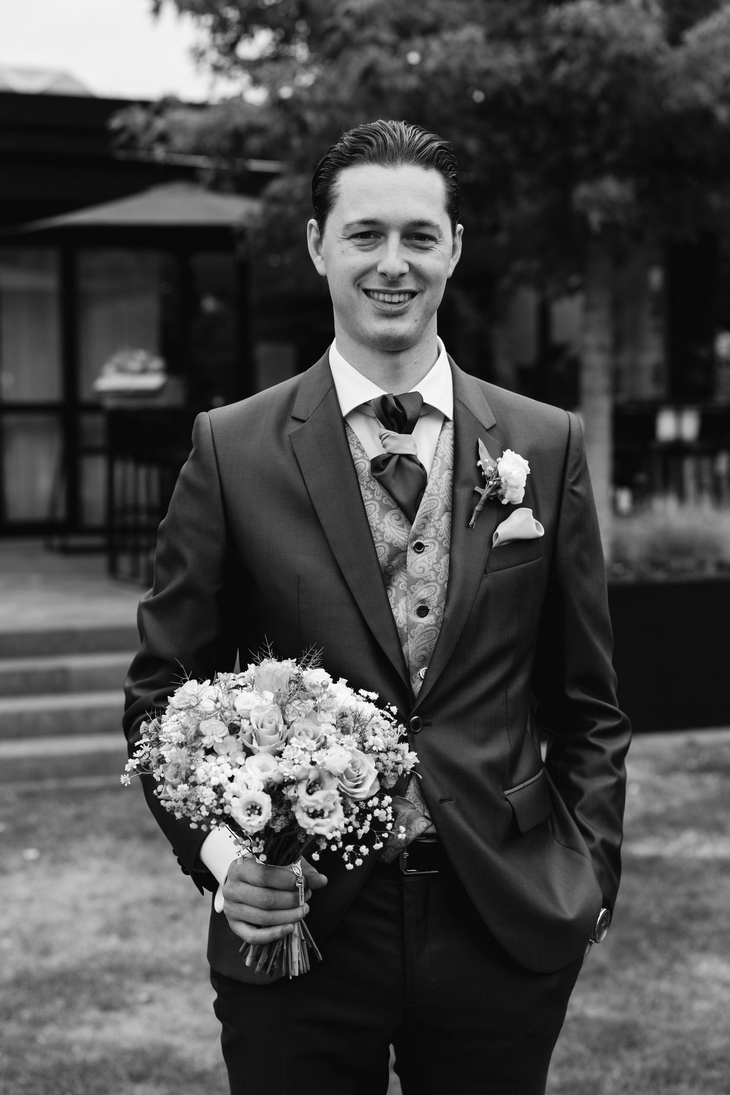 Groom in grey suit holding wedding bouquet smiling in black and white outdoor portrait.