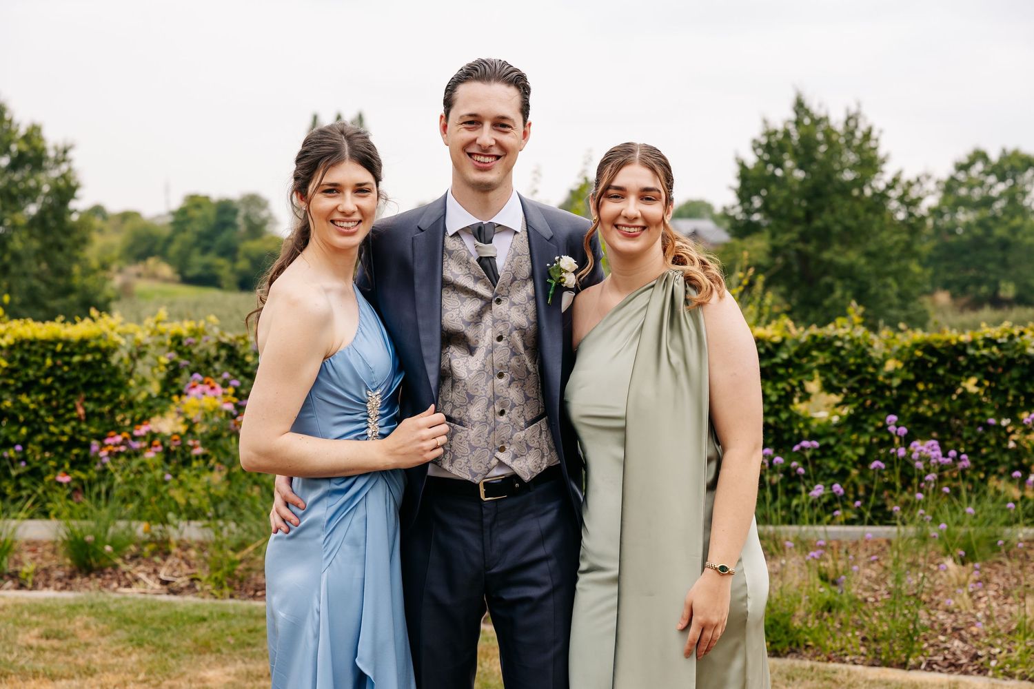 A smiling groom in a grey vest stands between two bridesmaids in blue and sage green dresses at a wedding.