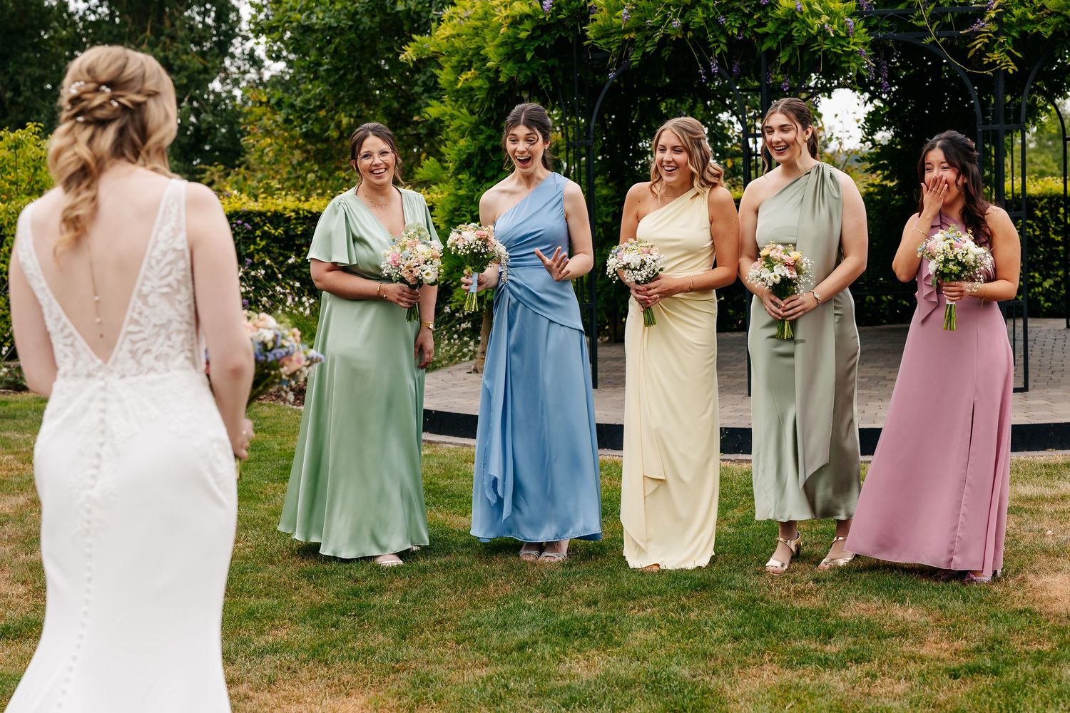 Bridesmaids in colorful pastel gowns smile at bride in white lace backless wedding dress in garden setting.