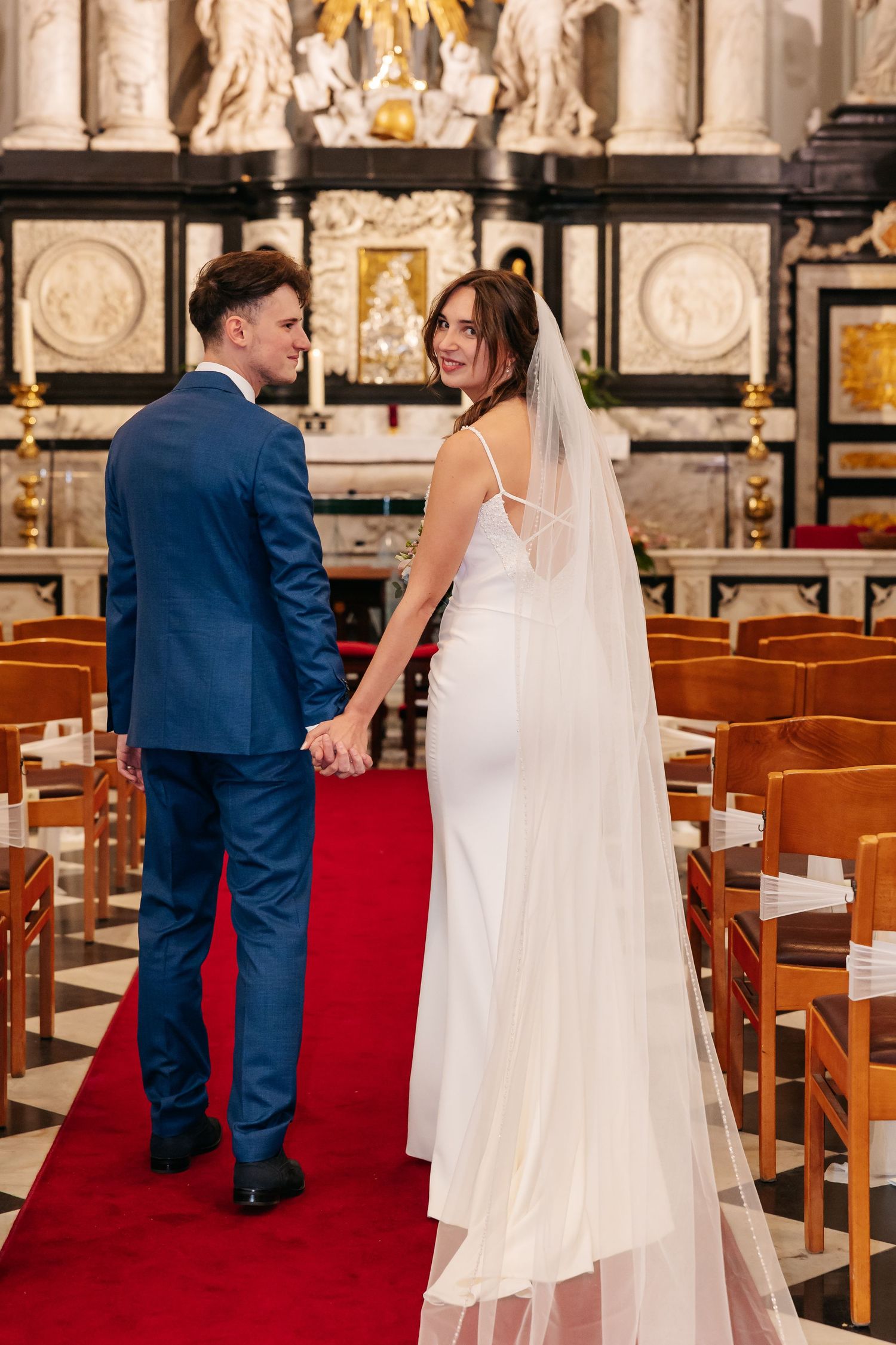 Smiling bride in white gown and veil walks with groom down church aisle.