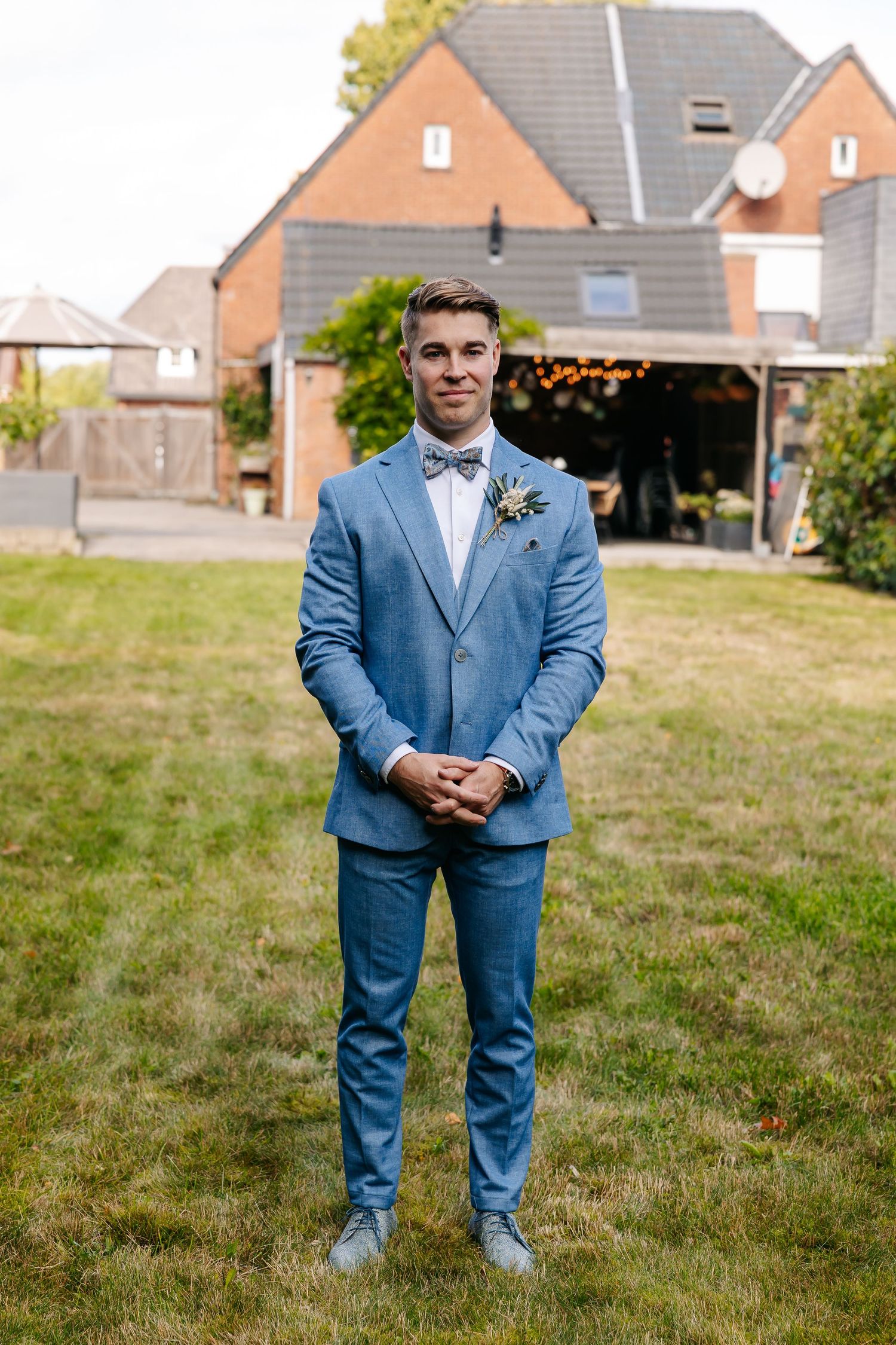 Groom in blue suit with bow tie and boutonniere standing on green lawn in front of brick house.