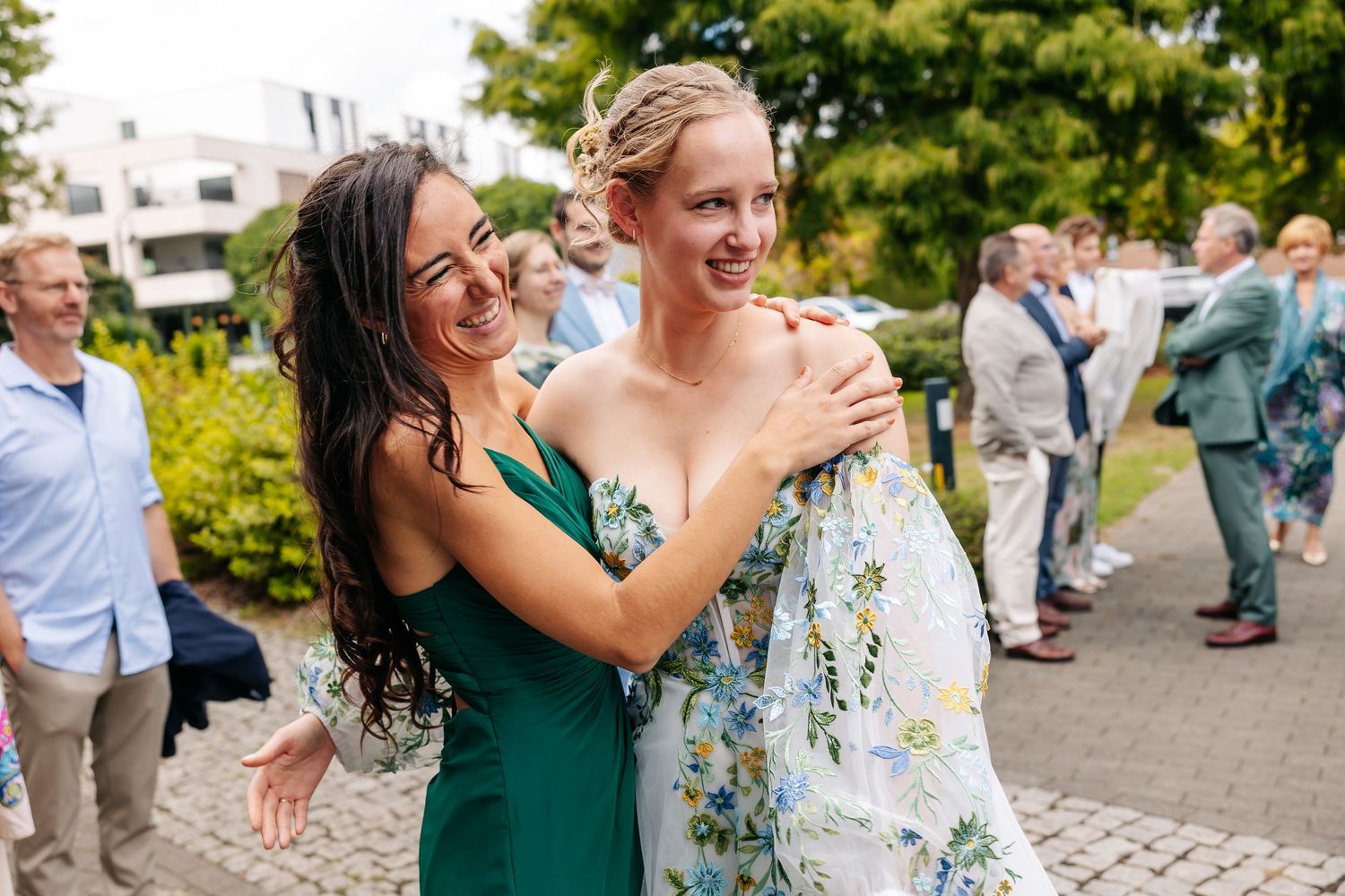 Two women embrace and smile at an outdoor wedding celebration, one in a green dress and one in a floral gown.