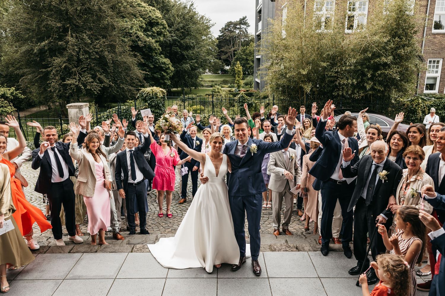 Newlyweds raise hands in celebration surrounded by cheering wedding guests outdoors.