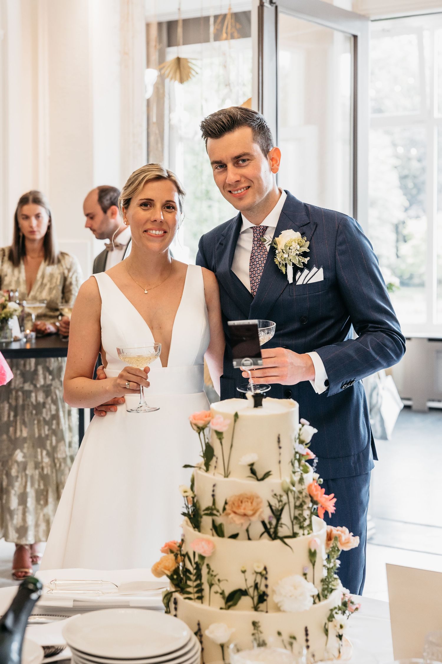 Smiling bride and groom posing beside floral wedding cake, holding champagne glasses at reception.