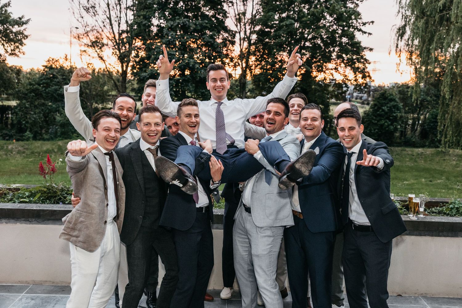 Groomsmen in suits celebrate by lifting groom outdoors at golden hour wedding reception.
