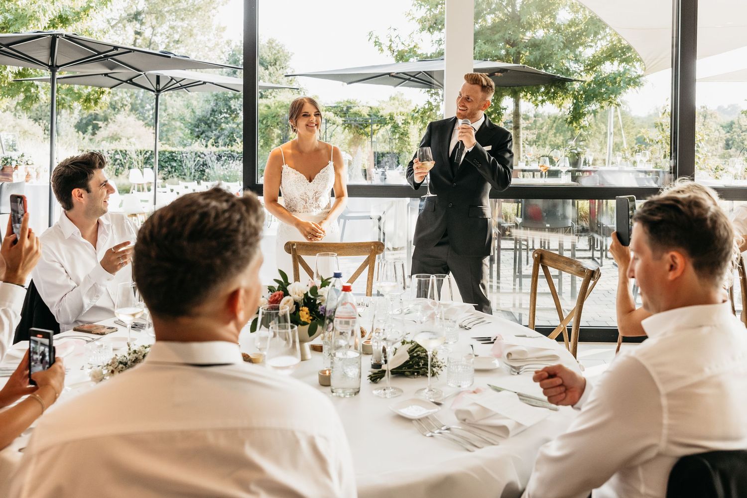 Groom giving a wedding speech while bride stands beside him at an elegant reception dinner.