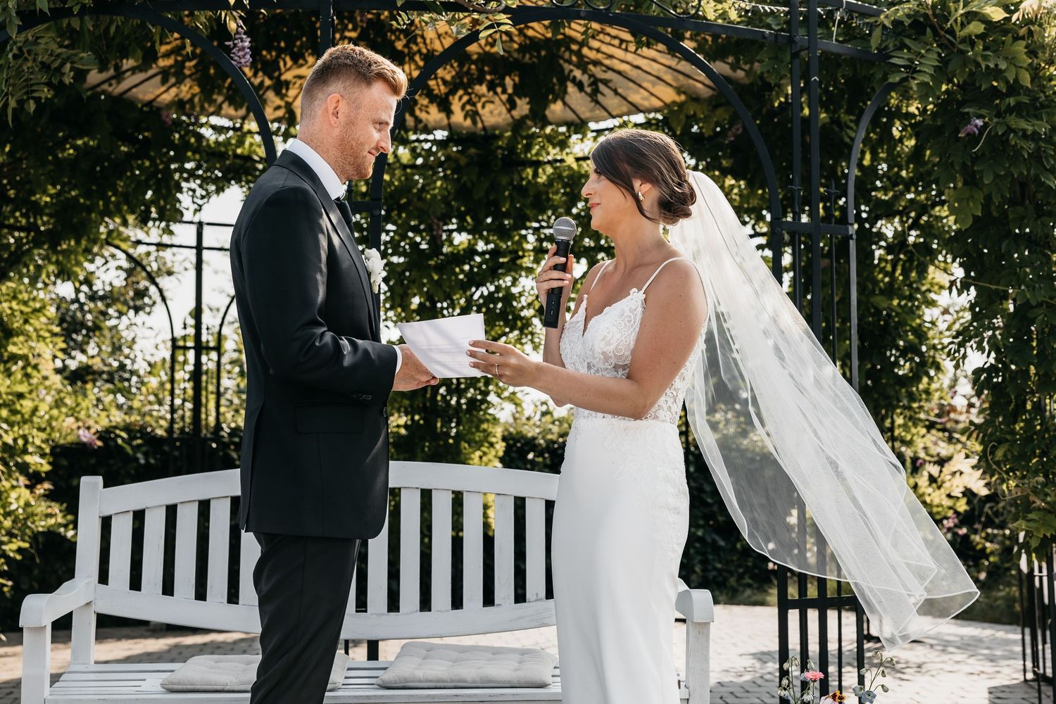 Bride reads heartfelt vows to groom beneath a lush garden arbor at outdoor ceremony.