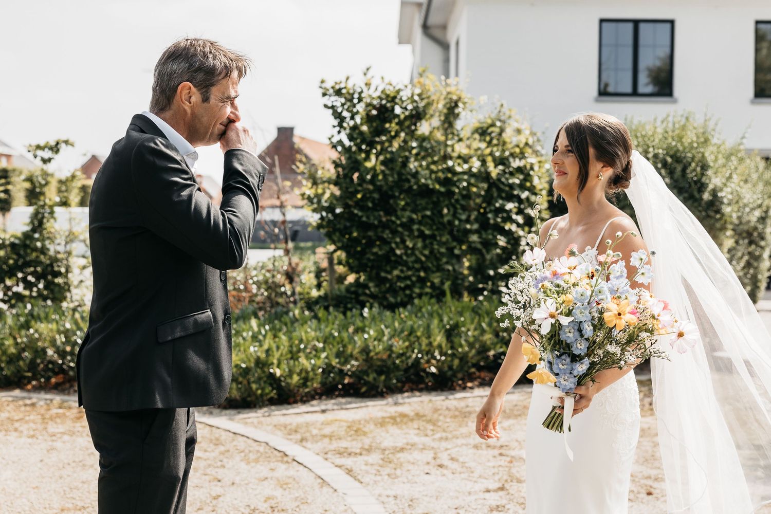 Emotional groom wiping tears seeing bride in white gown holding colorful bouquet during wedding first look outdoor.
