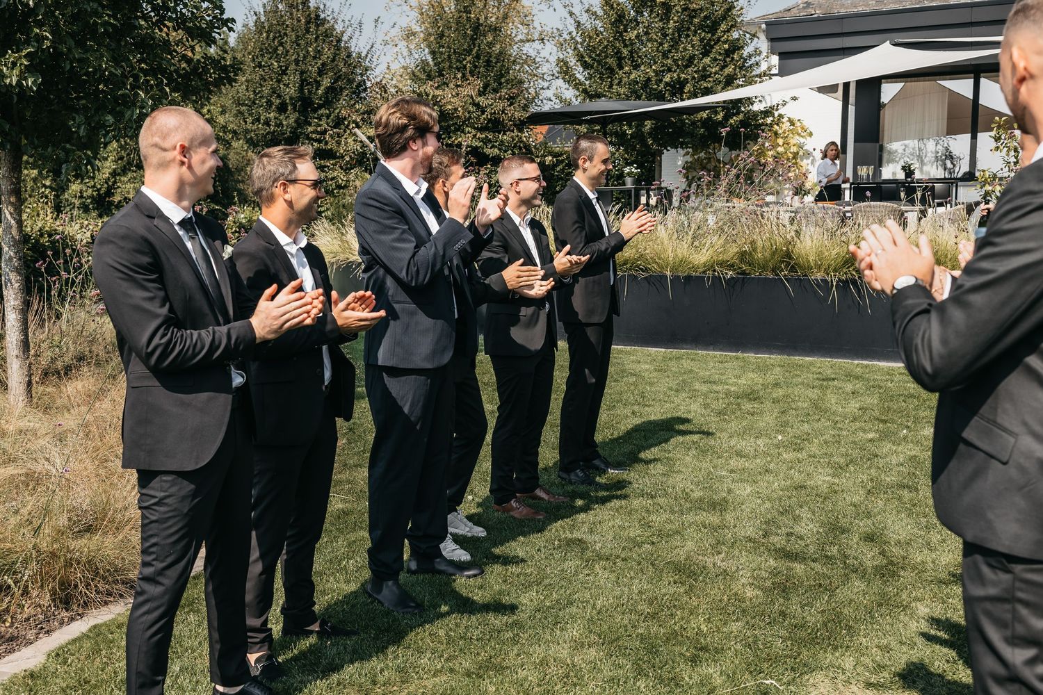 Groomsmen in black suits clapping outdoors at a wedding celebration on a sunny day.