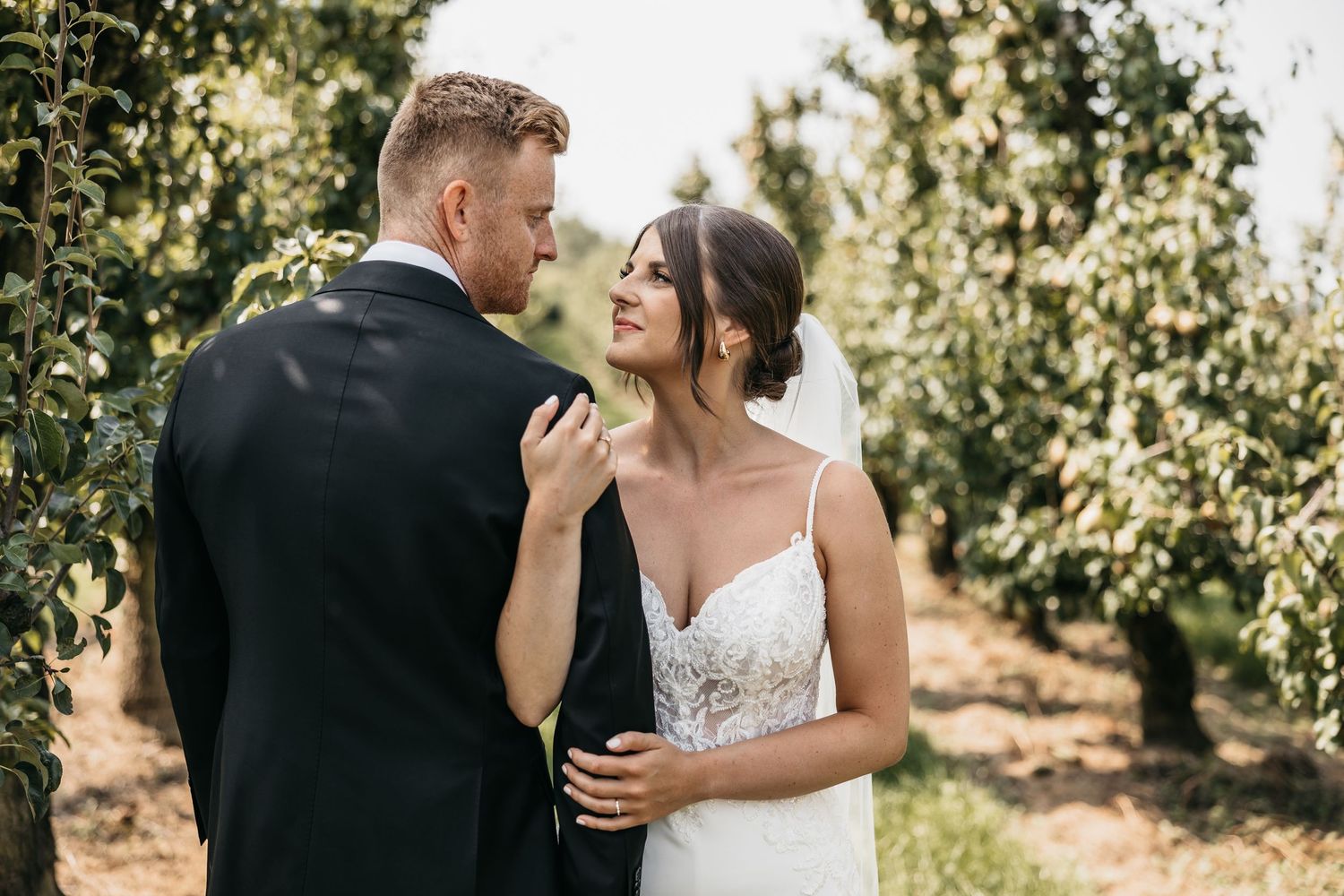 Bride smiles lovingly at groom while holding his lapel in a romantic orchard portrait.
