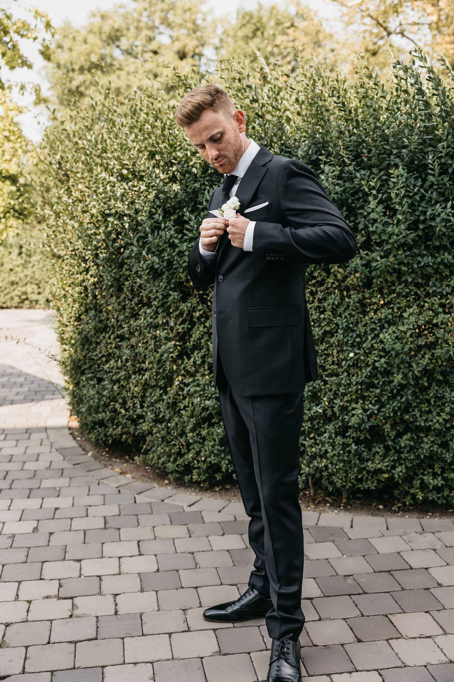 Groom in black tuxedo adjusting white boutonniere on lapel while standing on garden pathway.