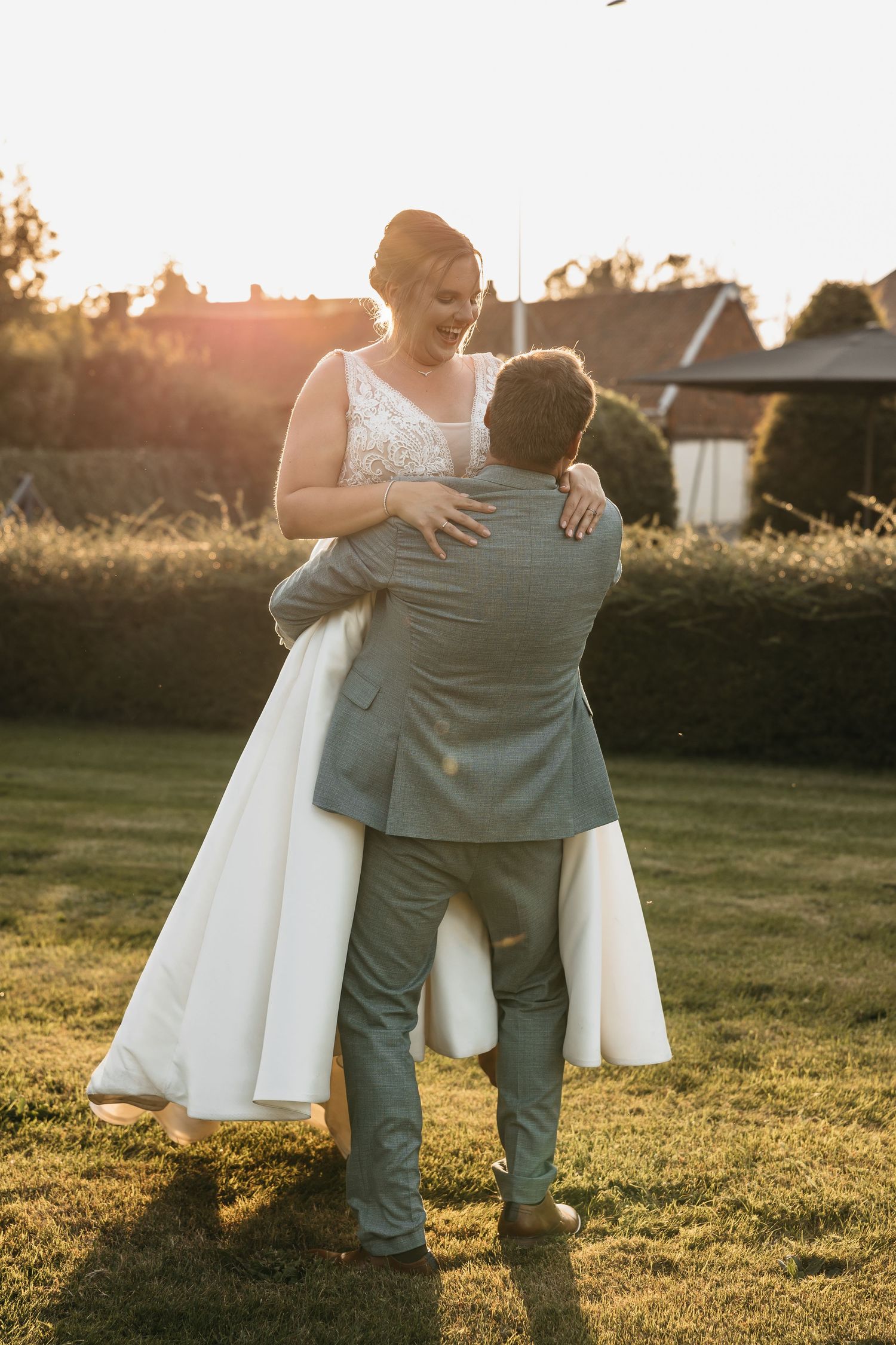 Groom lifting bride in lace wedding dress as she smiles in warm sunset glow.