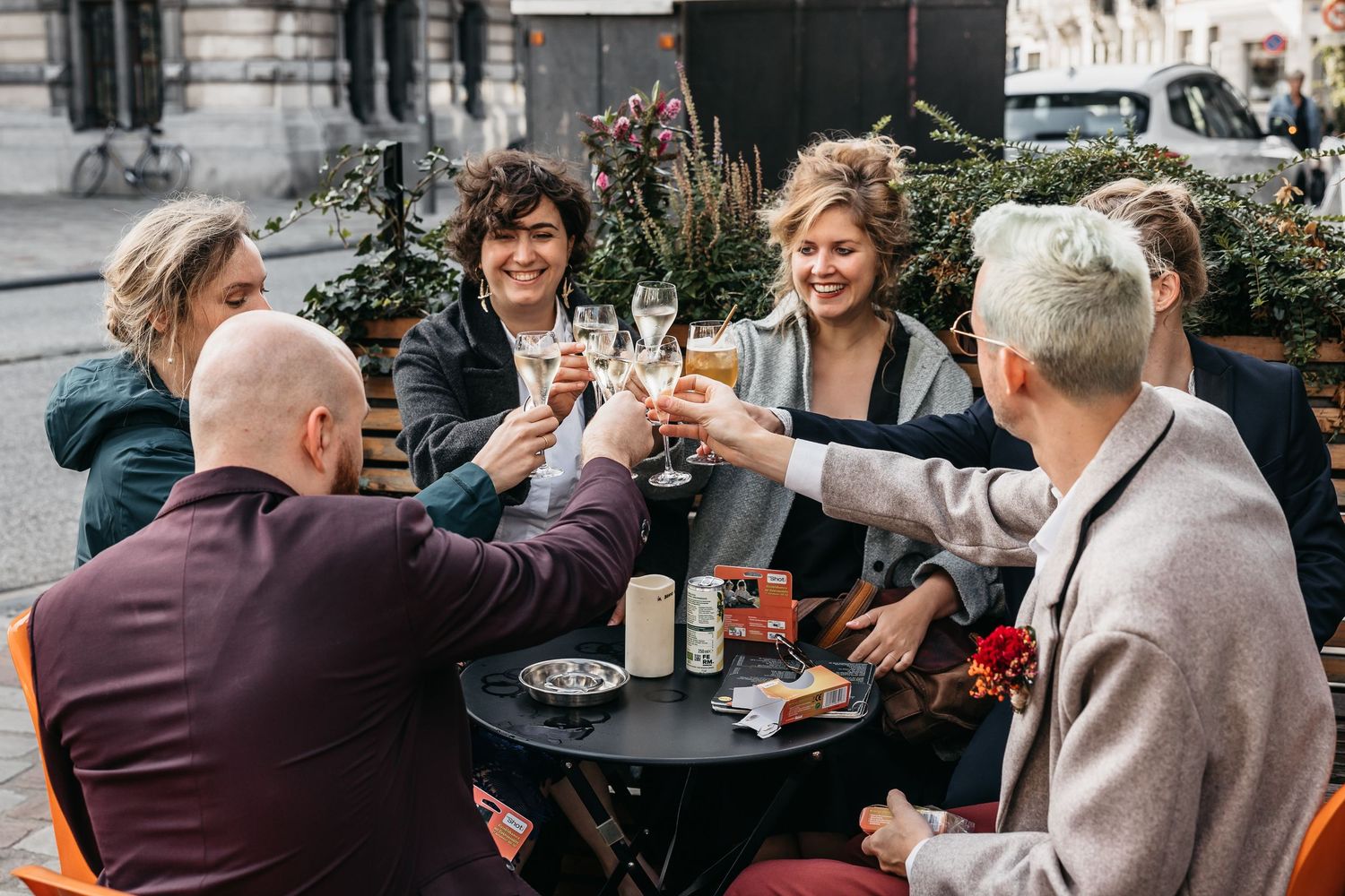 Group of six wedding guests toasting champagne glasses together at outdoor cafe table celebration.
