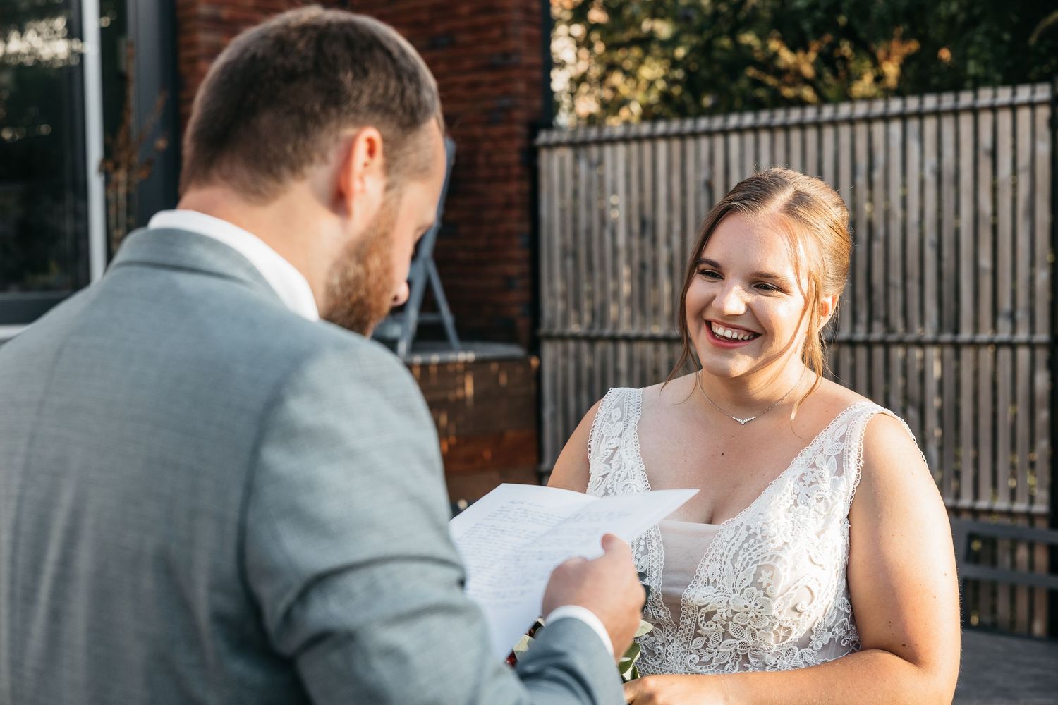Joyful bride in lace dress laughs as groom reads vows during ceremony.
