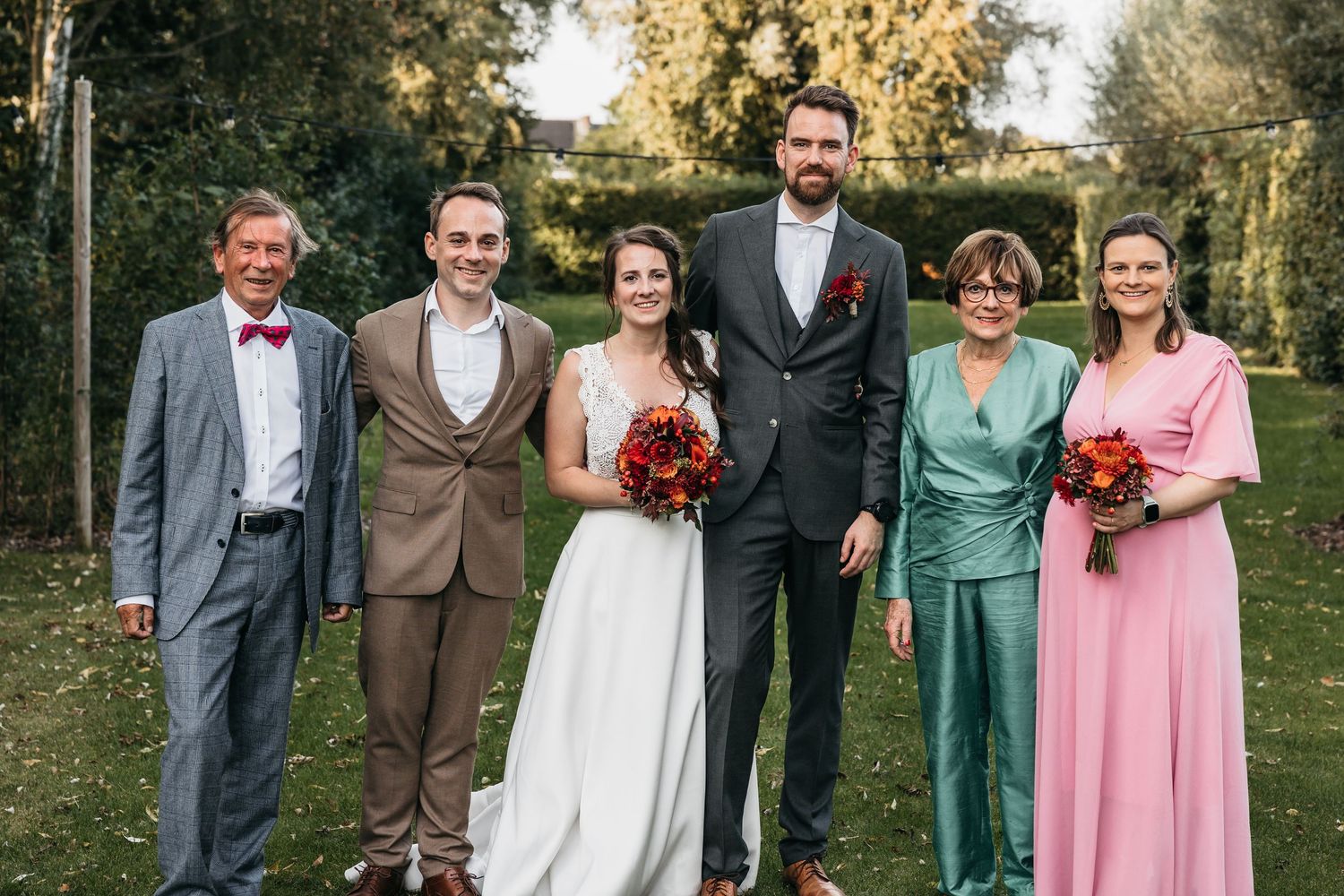 Bride and groom posing with four family members in garden setting with colorful formal attire and red bouquet.