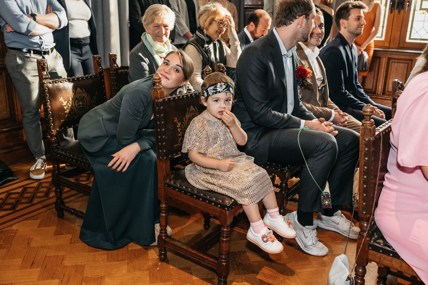 Woman in green dress crouching beside a young child in a hat seated at a wedding ceremony.