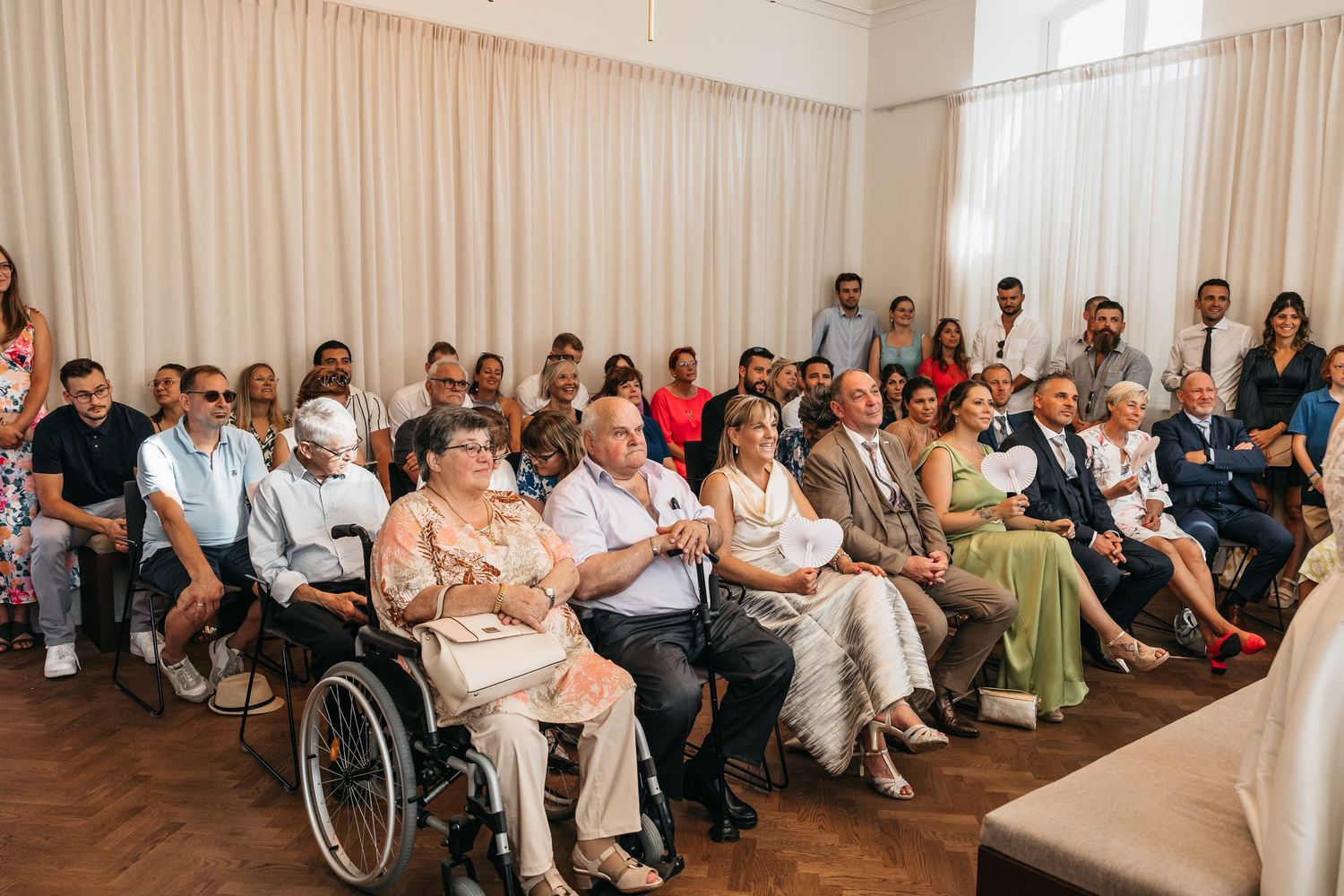 Wedding guests seated in rows including elderly person in wheelchair at indoor ceremony.