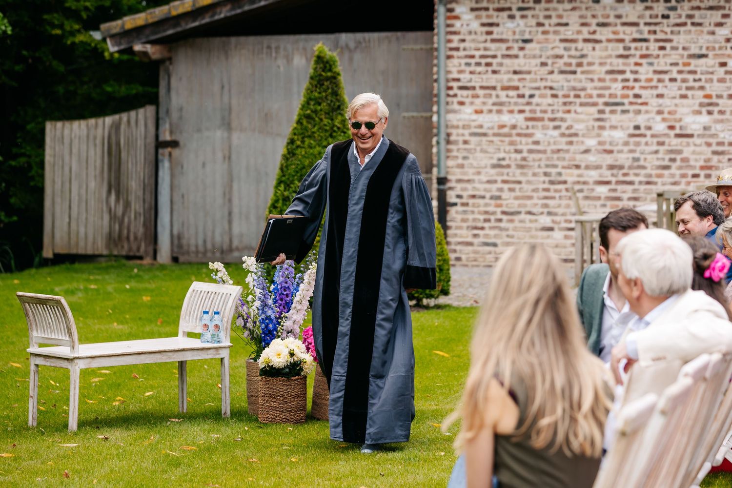 Officiant in black robe walking toward seated wedding guests at outdoor garden ceremony.