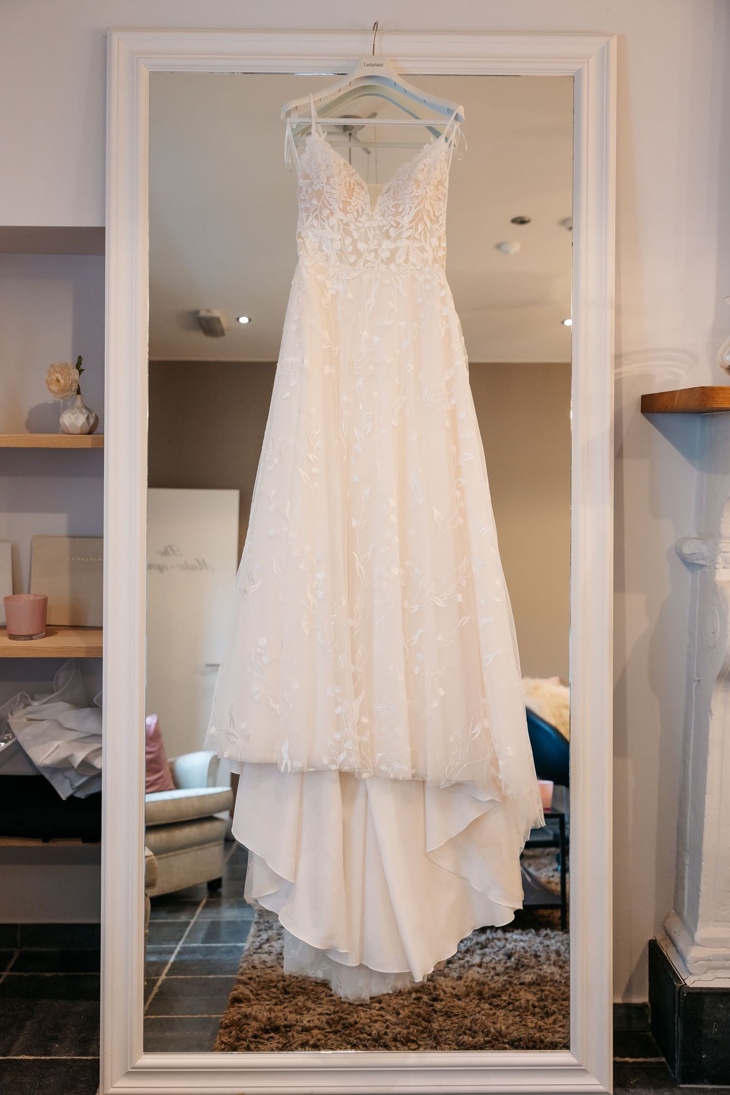 Elegant white lace wedding dress hanging on a hanger in a bridal boutique mirror reflection.